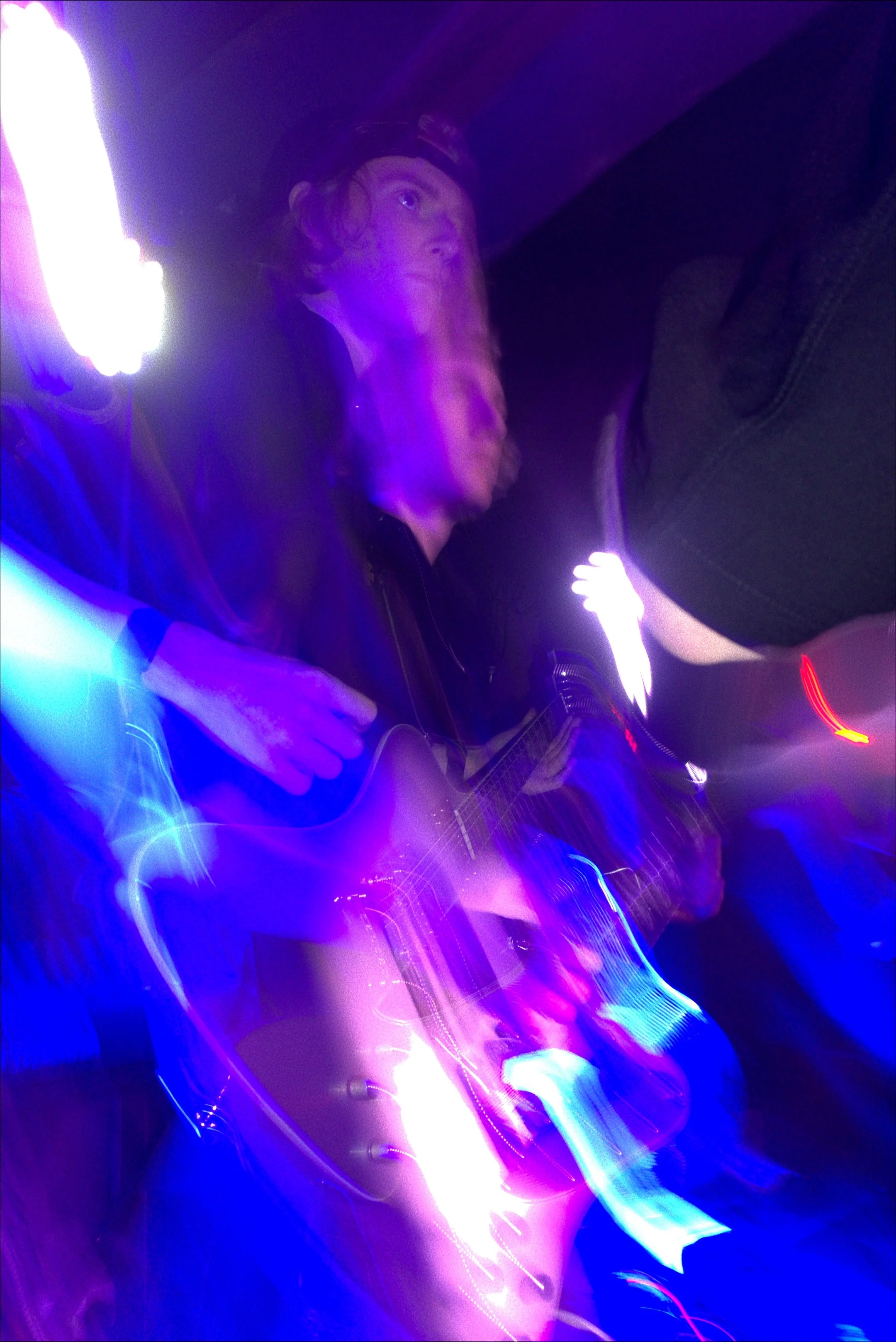A person with long hair wearing a headband, holding a guitar, surrounded by vibrant purple and blue neon lights in a dim environment.