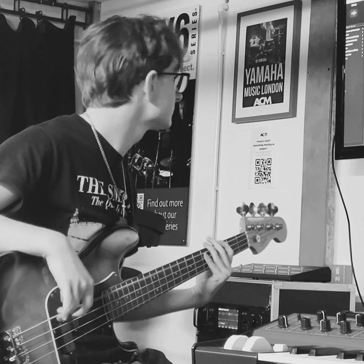A young man with glasses playing an acoustic guitar in a music studio with posters on the wall.