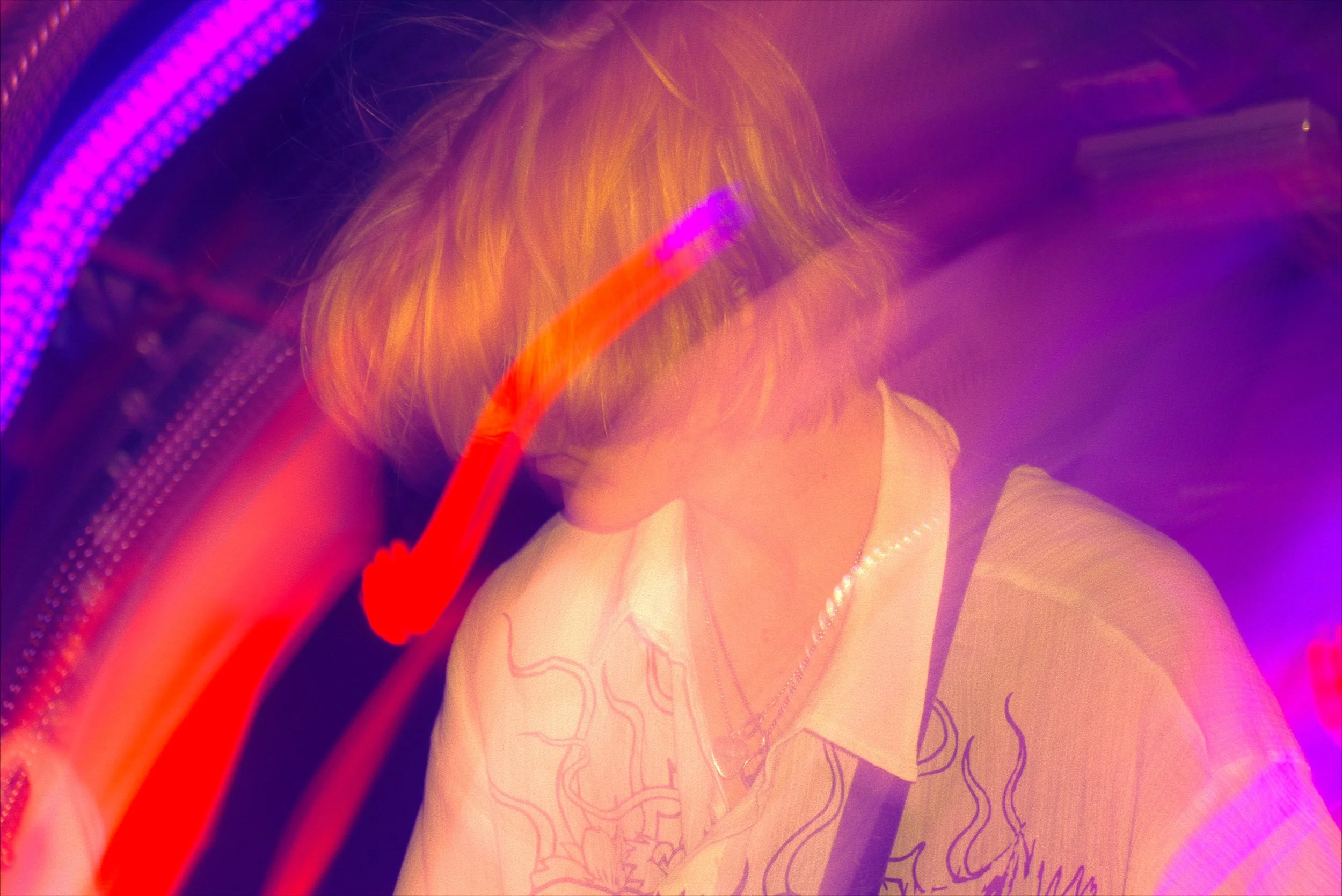 A person with blonde hair wearing a white shirt and a clear plastic bag on their shoulder, illuminated by purple and pink neon lights, with handprints and graphic designs on the shirt.