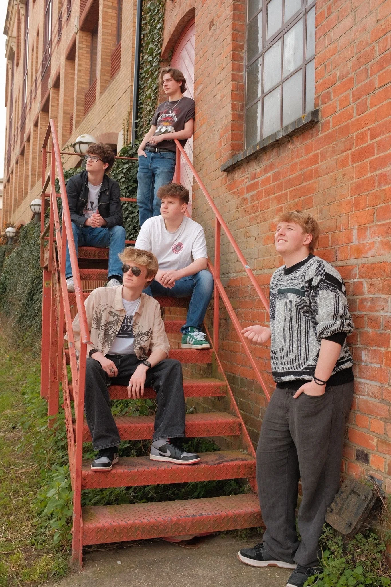A group of six young men on an outdoor metal staircase against a brick building, with one standing at the top and the others sitting or leaning on the stairs, some wearing sunglasses, posing for a photo in casual clothing.