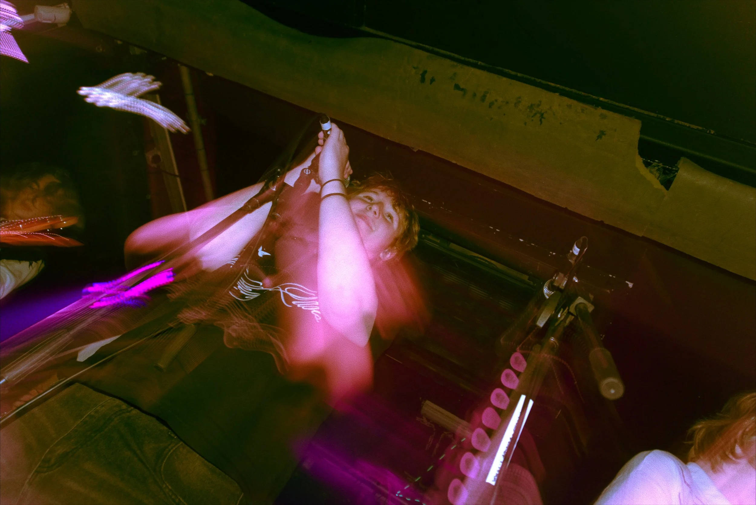A girl with curly red hair is lying on a bench, holding a guitar, with colorful, blurred light trails around her in a dimly lit environment.