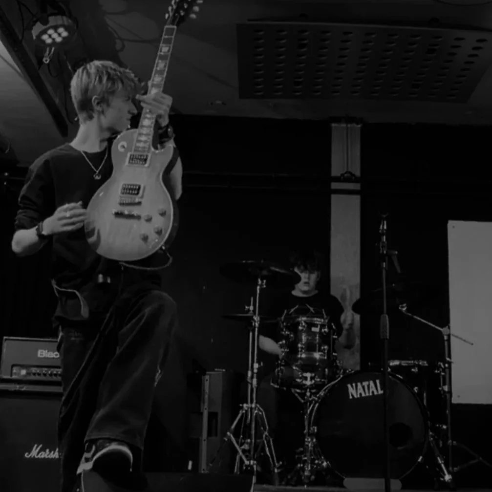 A young man playing an electric guitar on stage with a drummer in the background, in a music performance setting.