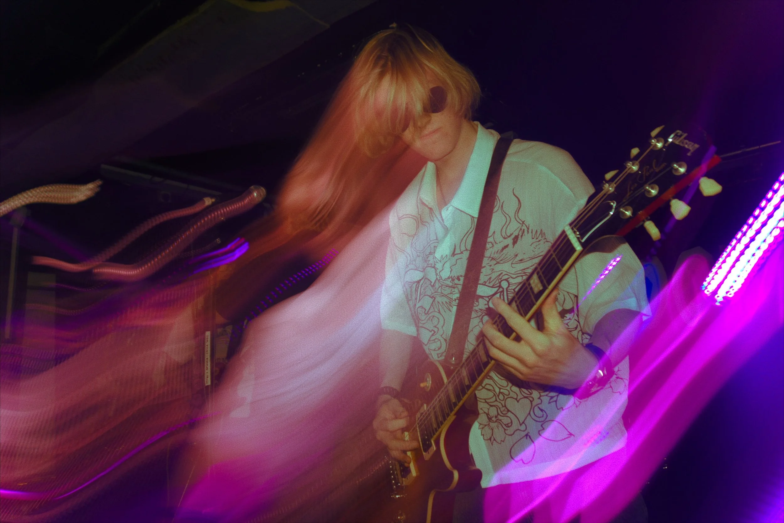 A person with long blonde hair playing an acoustic guitar on stage, wearing sunglasses and a white shirt with line art design, illuminated by purple stage lights.