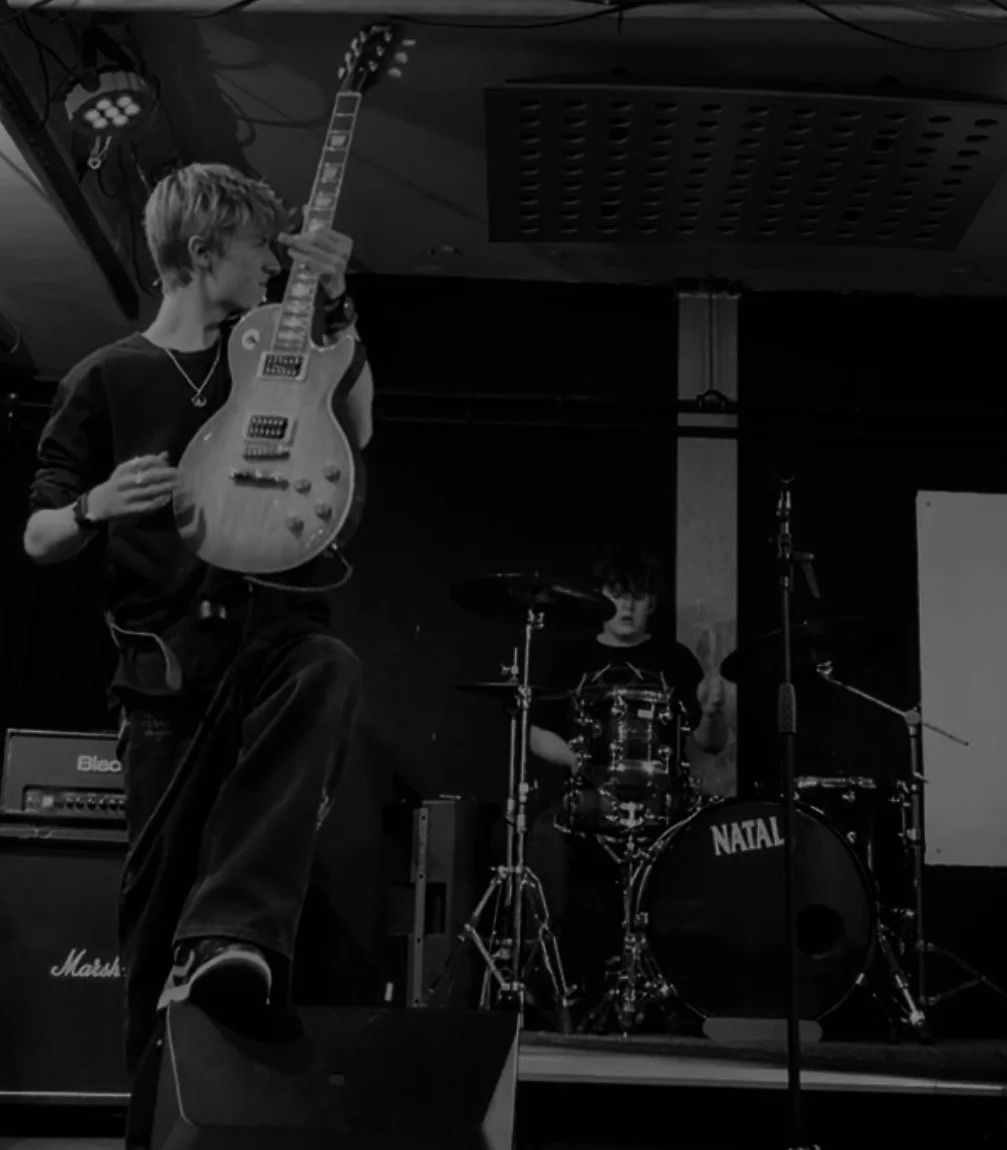 A young man playing an electric guitar on stage with a drummer in the background.