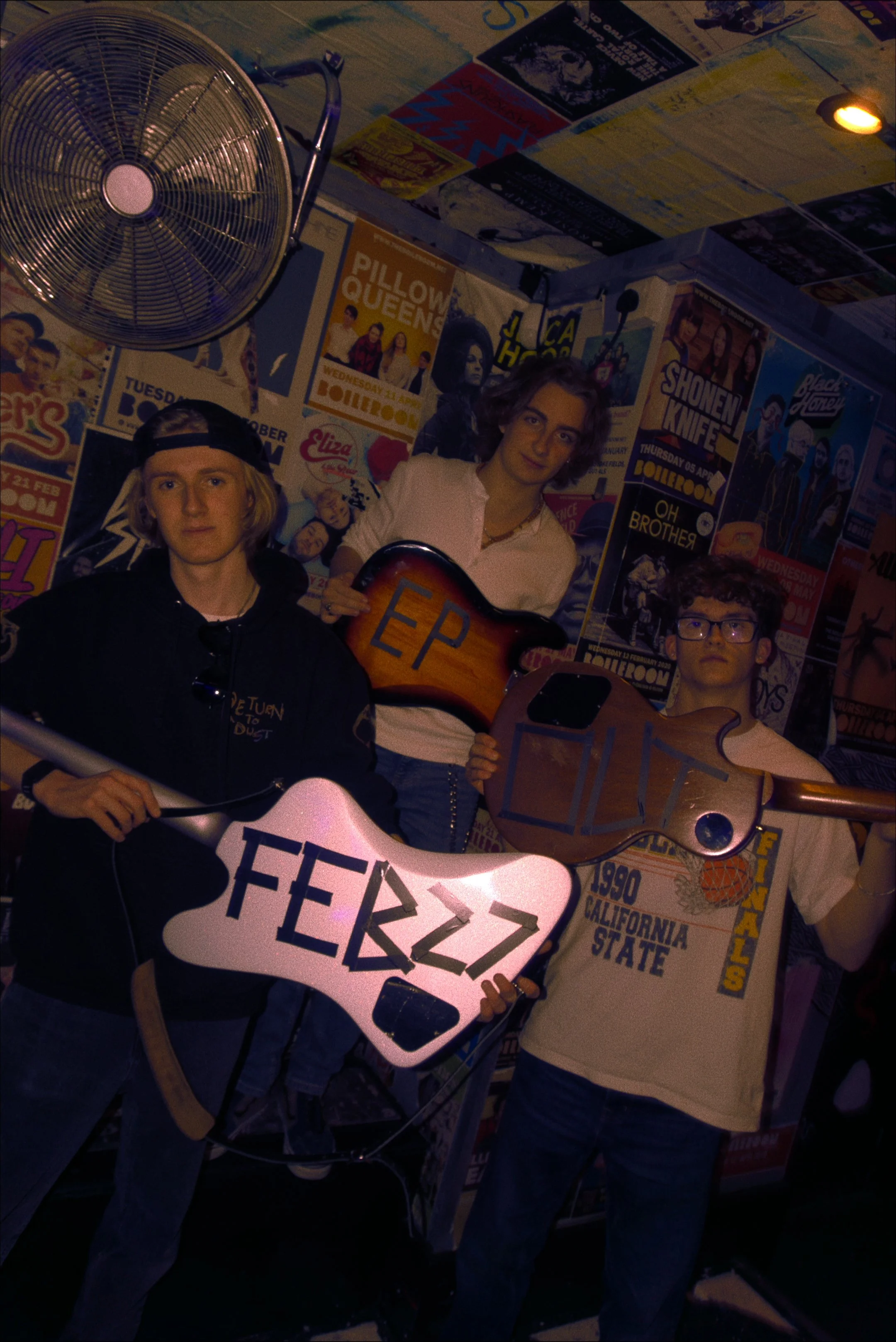 Three young men in a room with walls covered in posters, holding guitars and a sign with some Greek letters. One is wearing a black hoodie and a backwards baseball cap, the second is wearing a white shirt and holding an electric guitar, and the third