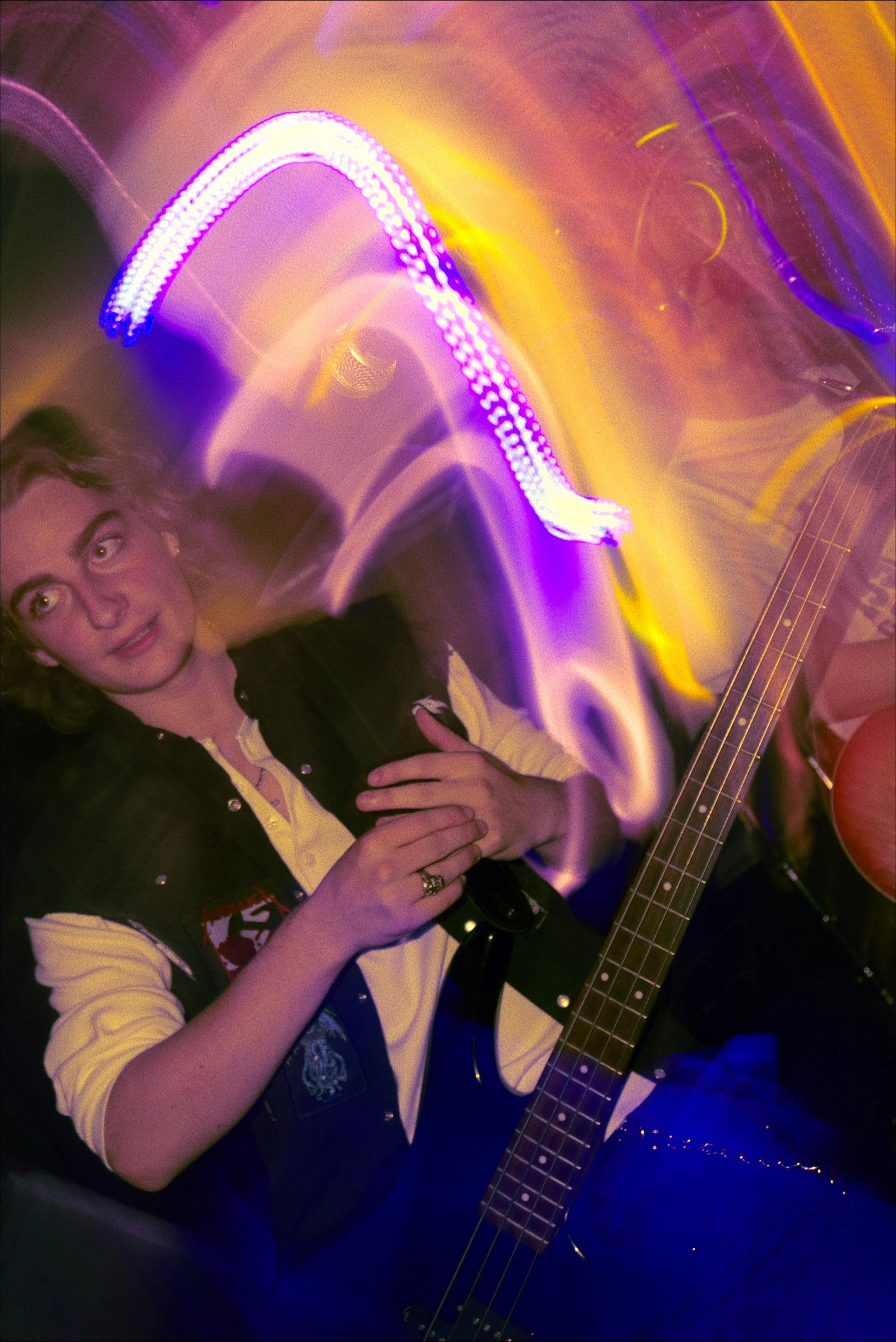 A young woman with long hair and a vest over a yellow shirt is holding a guitar. She is standing in a room with colorful, blurred lights, possibly at a party or a concert.