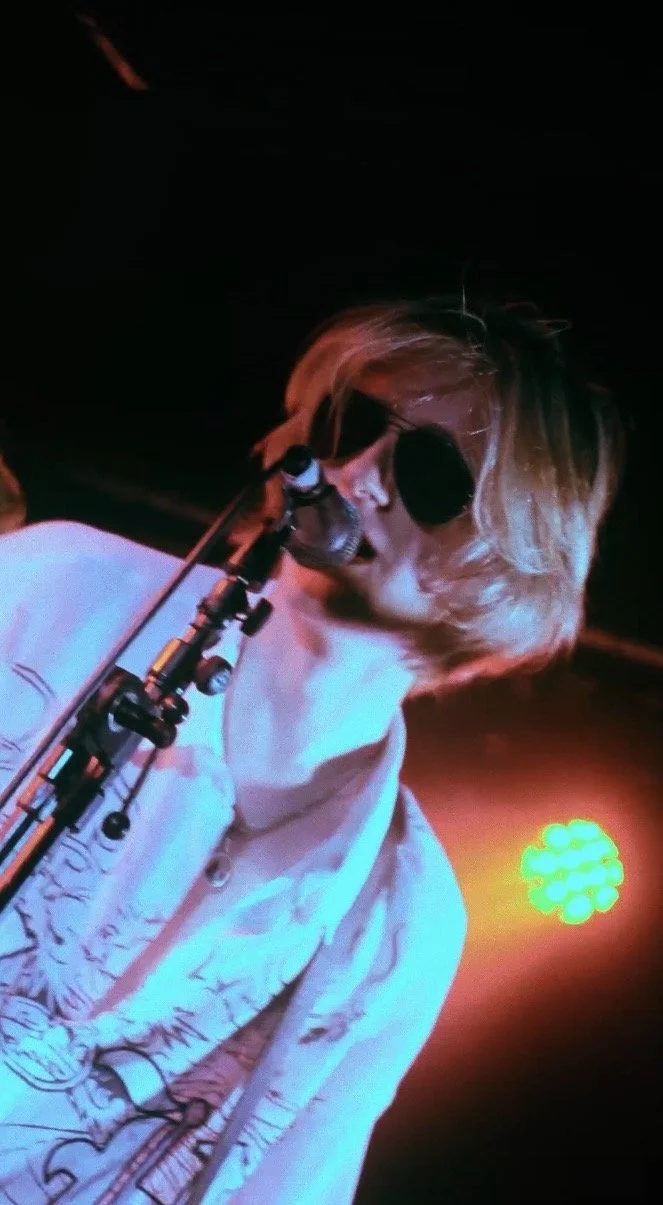 A woman with blonde hair, wearing sunglasses and a light-colored jacket, singing into a microphone on stage with colored stage lights behind her.