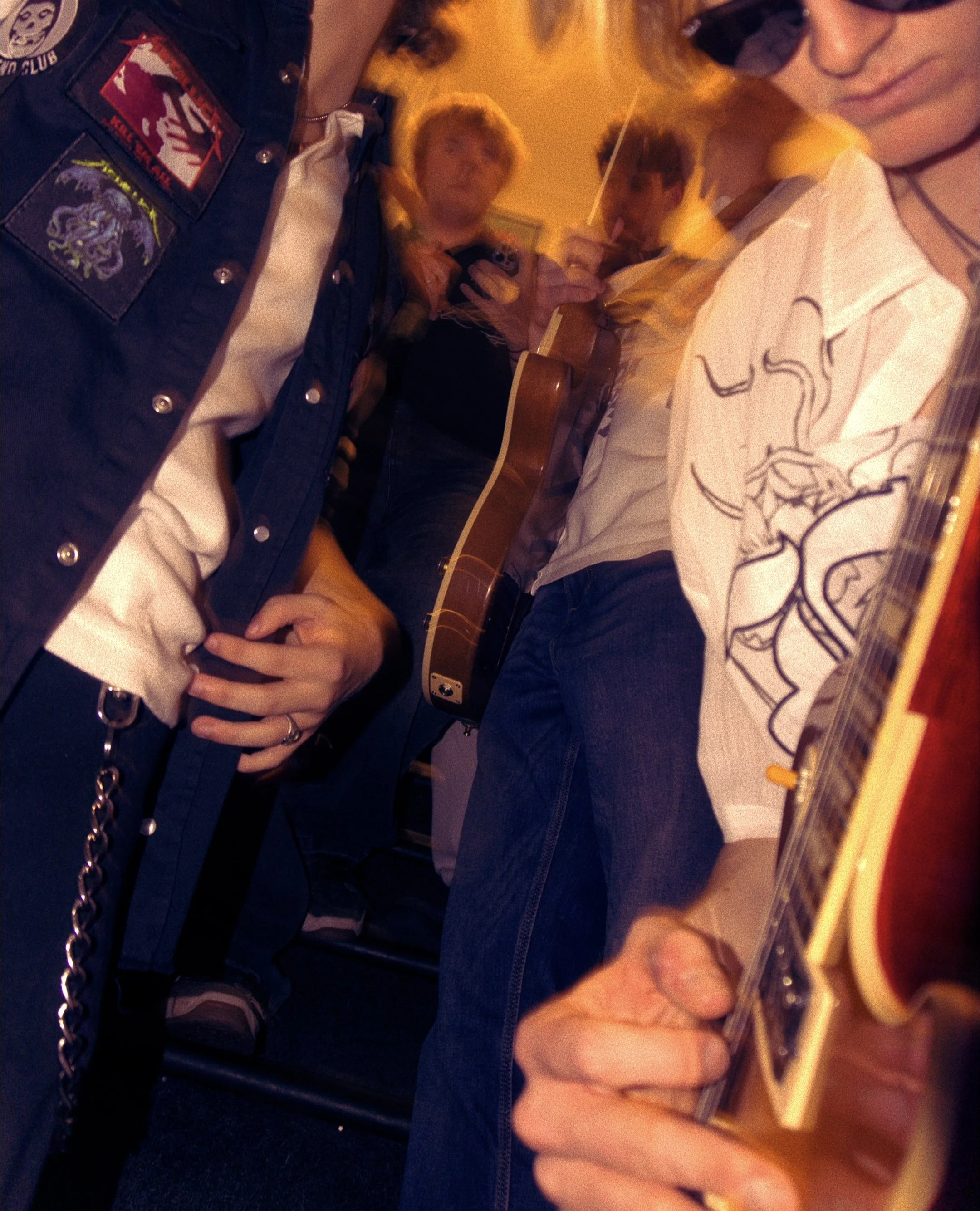Close-up of a person playing an acoustic guitar with a decorated body, wearing a white shirt with black outlines of animal drawings. Some people are visible in the blurred background, including a person with a guitar and someone holding a phone. The 