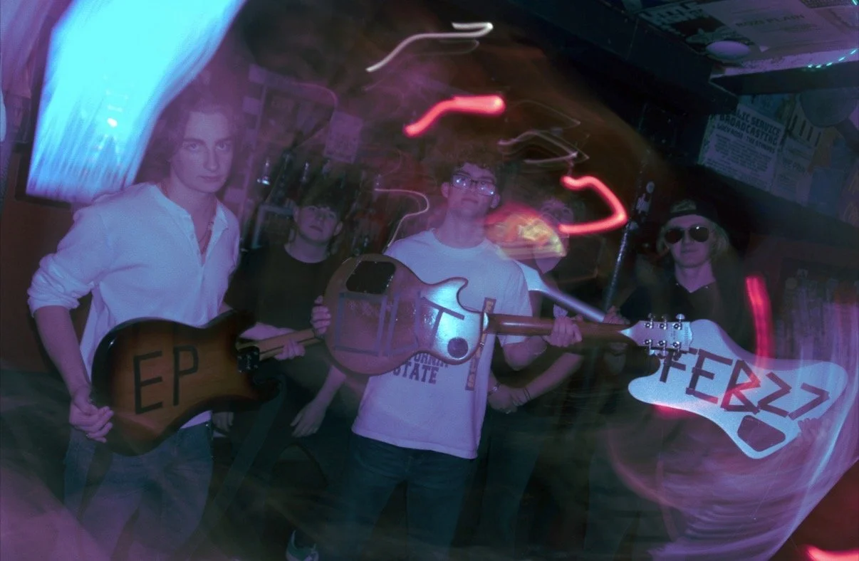 Four people with guitars in a dimly lit room with purple and red neon lights, blurry motion effects, and posters on the wall.