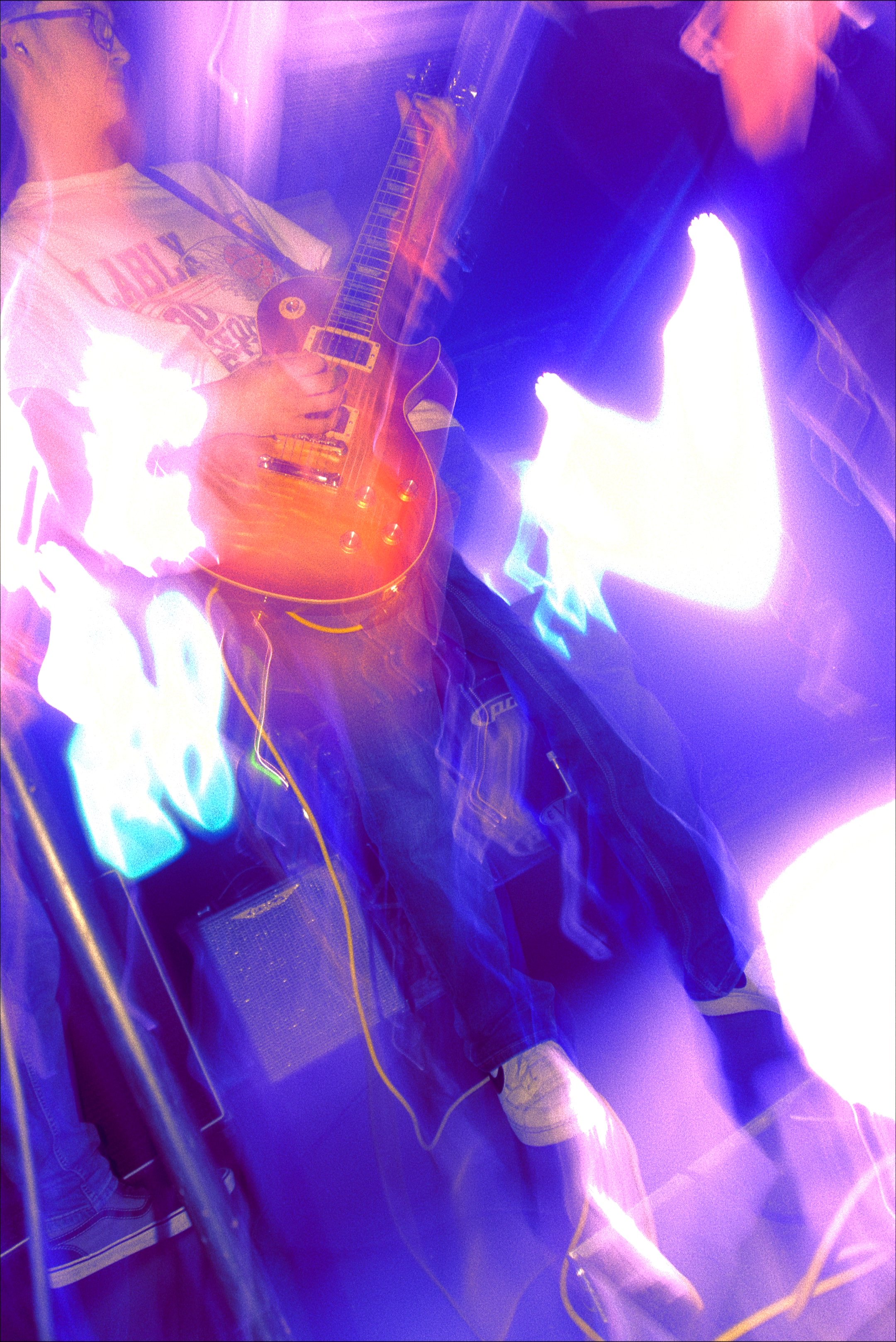 Two young men are in an elevator, with one playing an electric guitar and the other wearing glasses. The photo has a vibrant, colorful, and abstract lighting effect with strong purple, pink, and blue hues.
