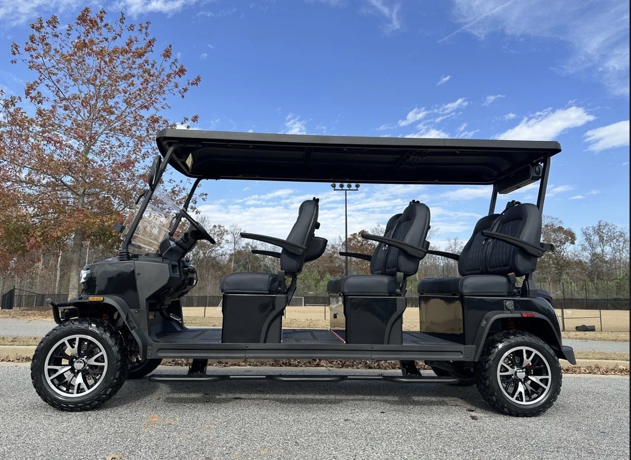Black golf cart with four seats parked on a paved surface with trees and a sports field in the background under a partly cloudy sky.