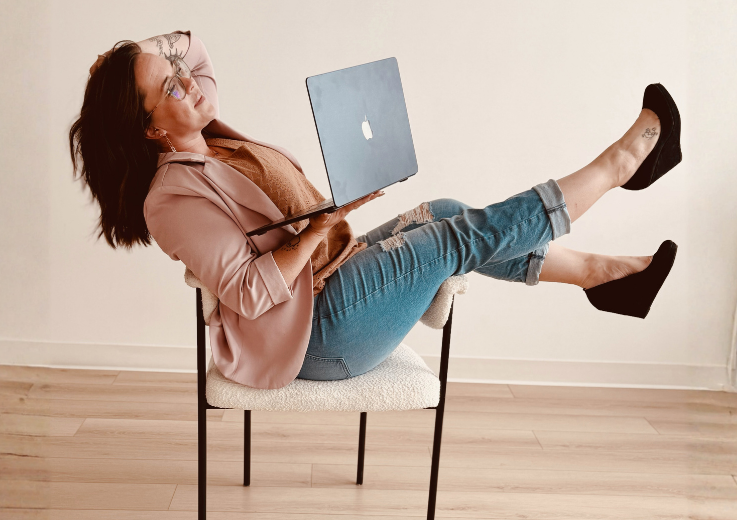 Woman sitting on a chair with her legs extended, holding a laptop, wearing glasses, a pink blazer, ripped jeans, and black shoes, against a plain white wall.