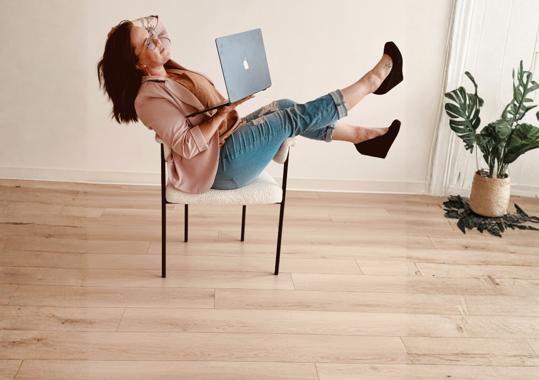 Woman sitting on a chair with legs up, holding a laptop, in a room with wooden floor and a potted plant.