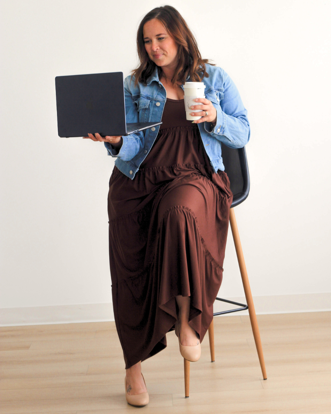 Woman sitting on a stool, holding a laptop in her left hand and a disposable coffee cup in her right hand, wearing a denim jacket, brown dress, and beige high heels.