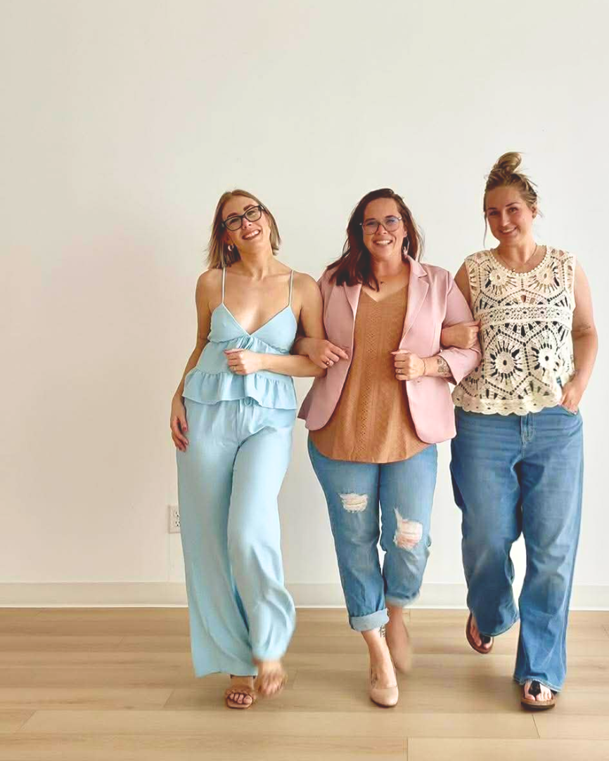 Three women walking arm in arm, smiling, in casual clothing, in front of a plain wall.