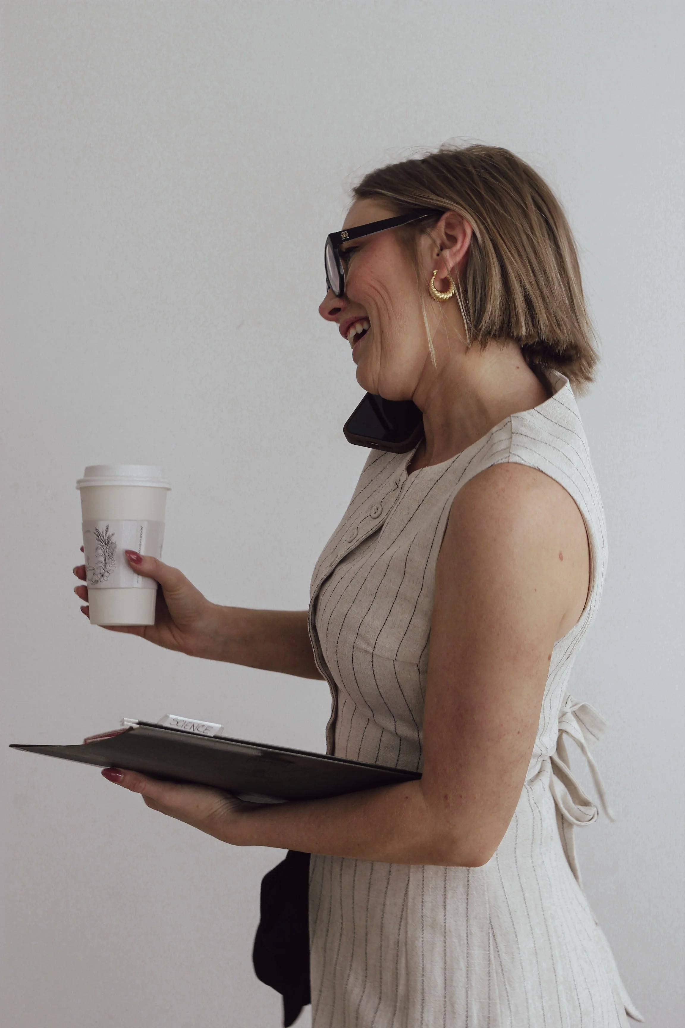 A woman with shoulder-length brown hair, wearing glasses and gold hoop earrings, smiling while holding a disposable coffee cup in her right hand and a clipboard in her left hand. She is dressed in a sleeveless beige pinstriped dress with a bow at the back.