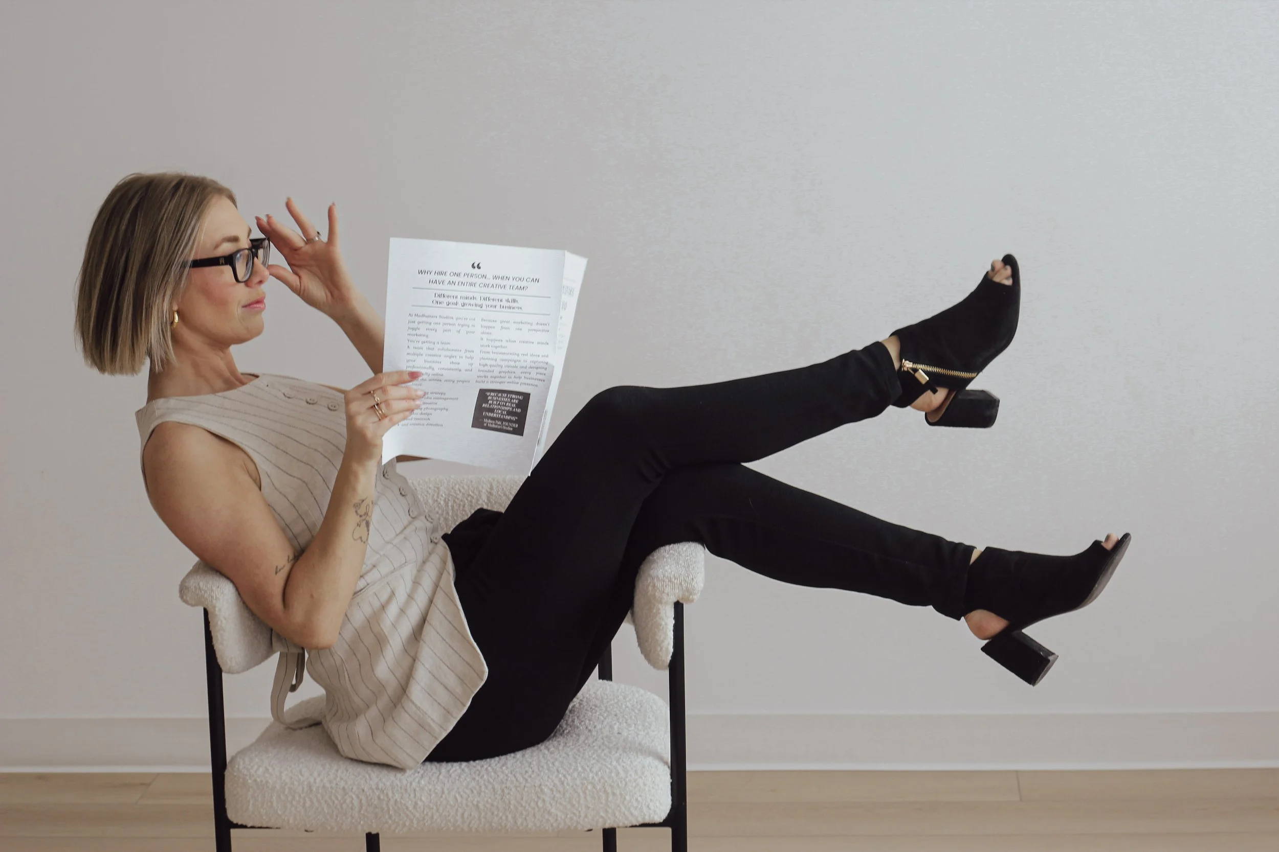 A woman with short blonde hair, wearing glasses, a sleeveless beige striped top, and black pants, is sitting on a chair with her legs raised and crossed at the ankles. She is reading a printed document and has a neutral expression. The background is plain light gray, and the floor is wooden.