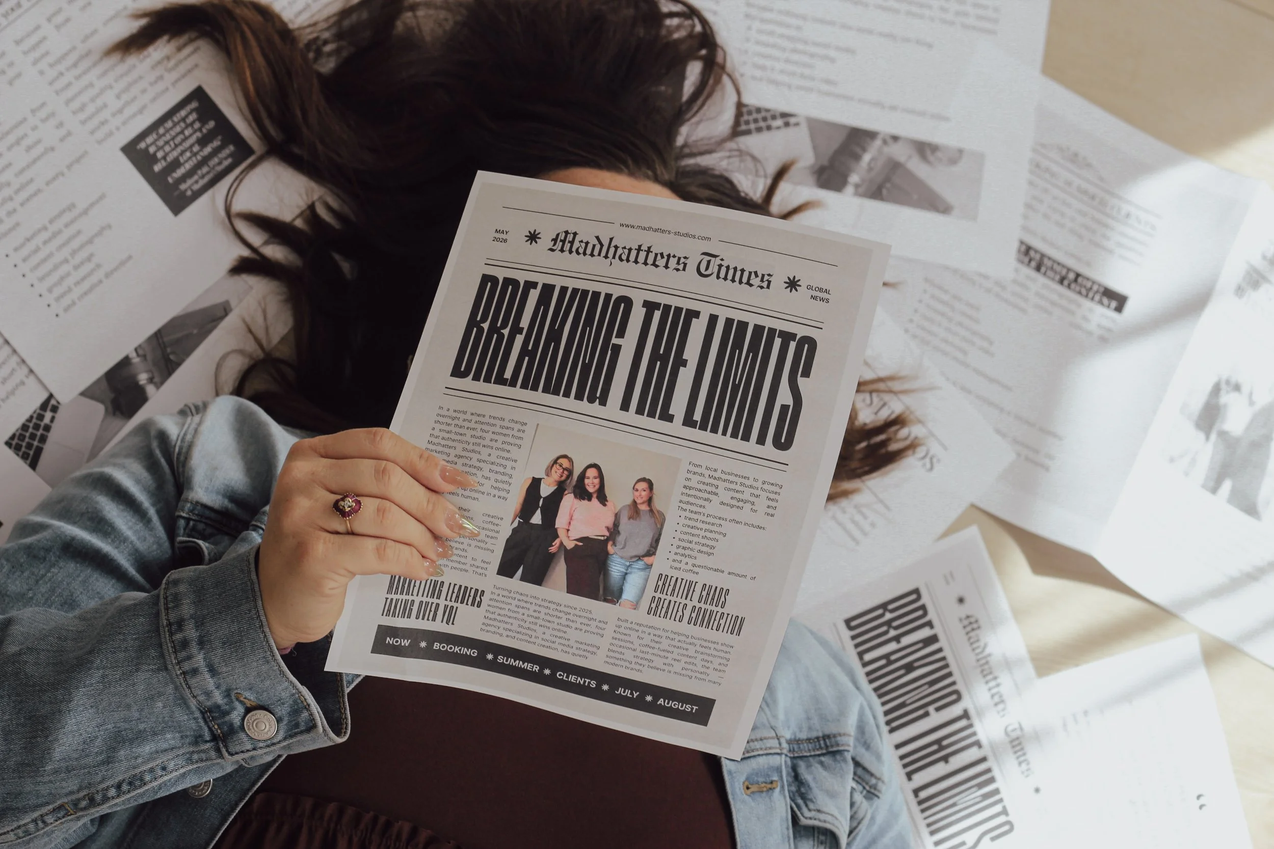 A person holding a newspaper titled "Madhatters Times" with the headline "BREAKING THE LIMITS". Other newspapers and printed sheets are scattered around them on a surface.