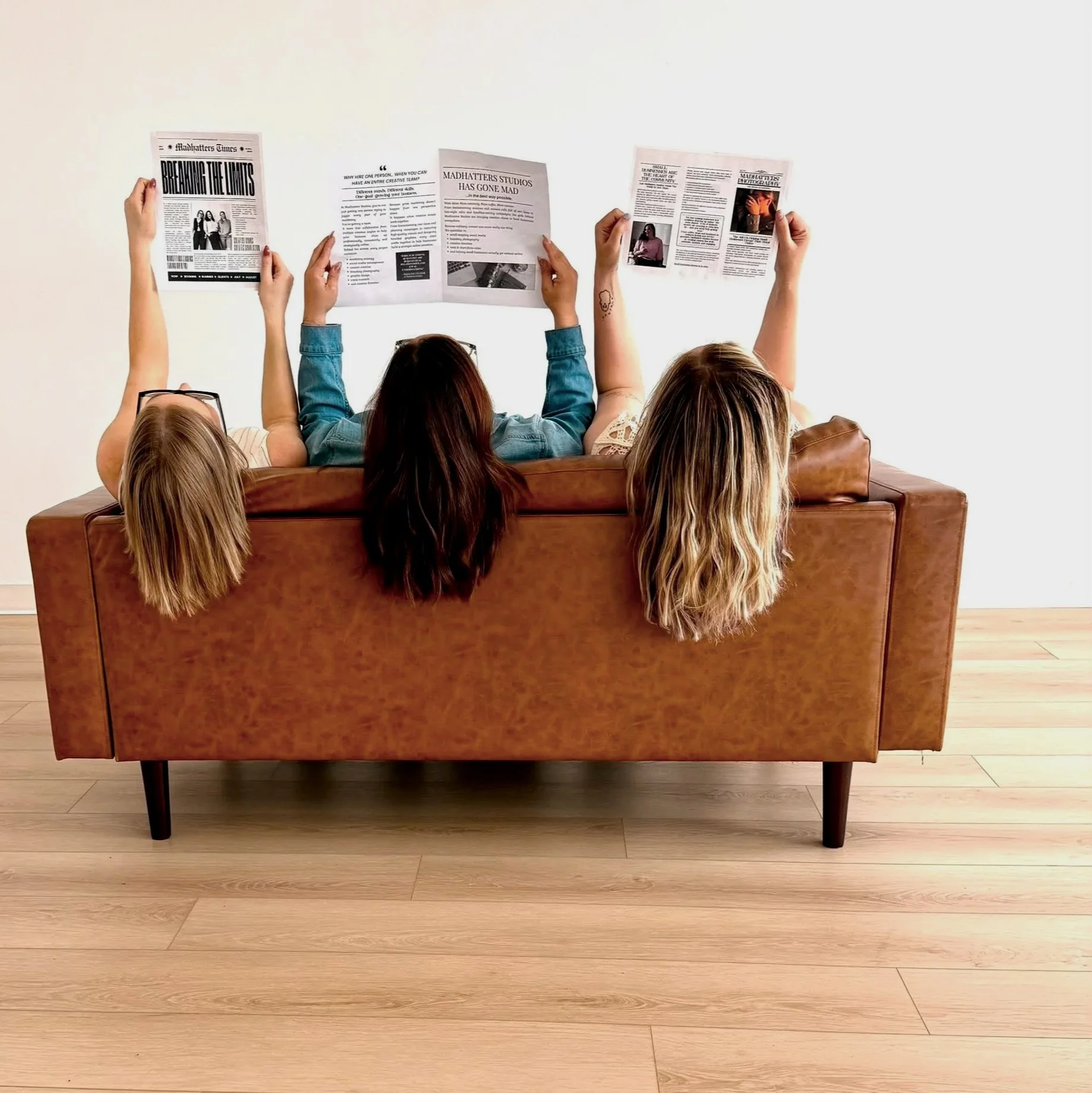 People sitting on a brown sofa reading newspapers, with light-colored wooden flooring.