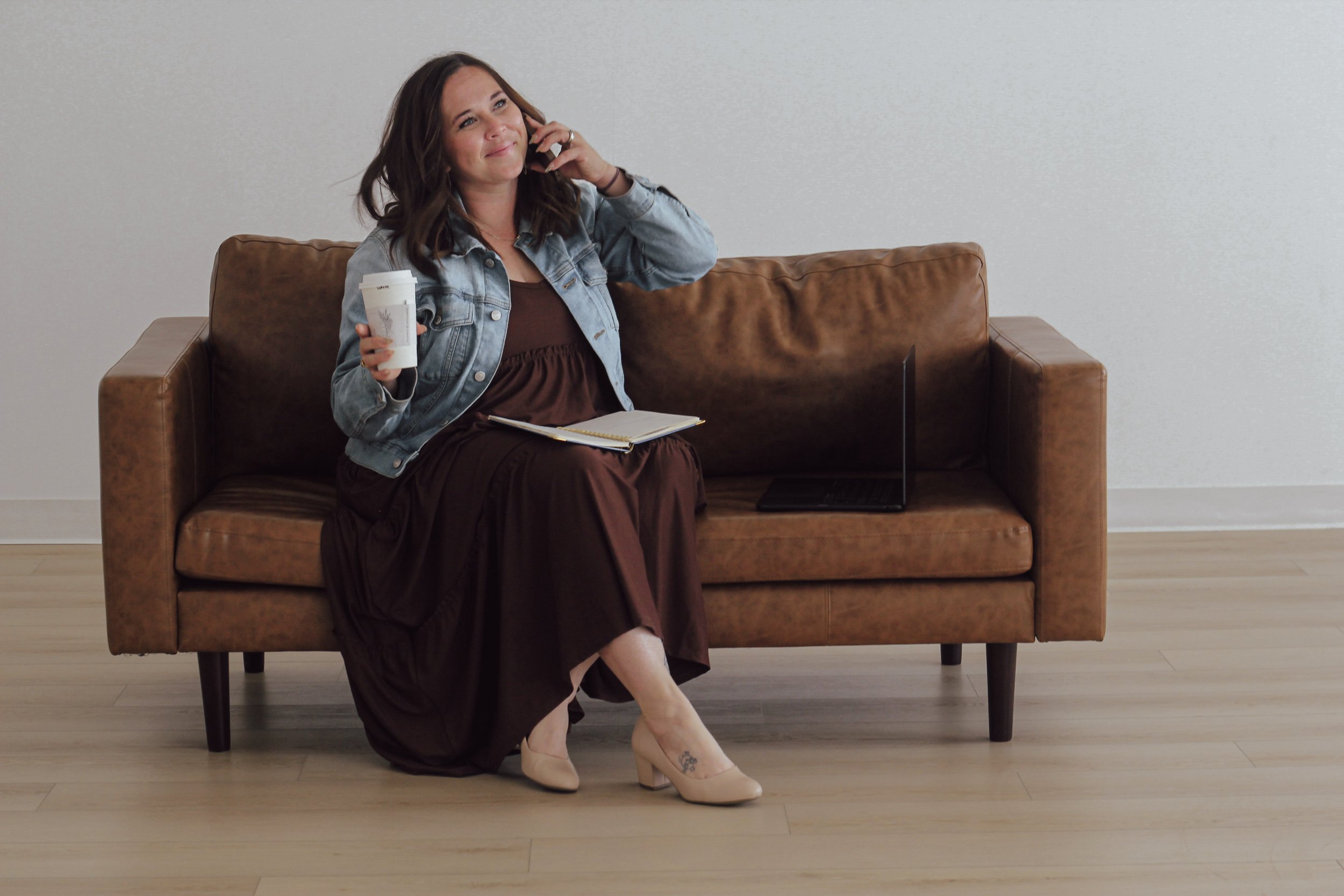 A woman is sitting on a brown leather couch, talking on her cellphone while holding a takeaway coffee cup in her right hand. She is wearing a denim jacket, brown dress, and beige high heels, with a notebook on her lap. An open laptop is placed on the couch beside her. The background features a plain, light-colored wall and wooden flooring.