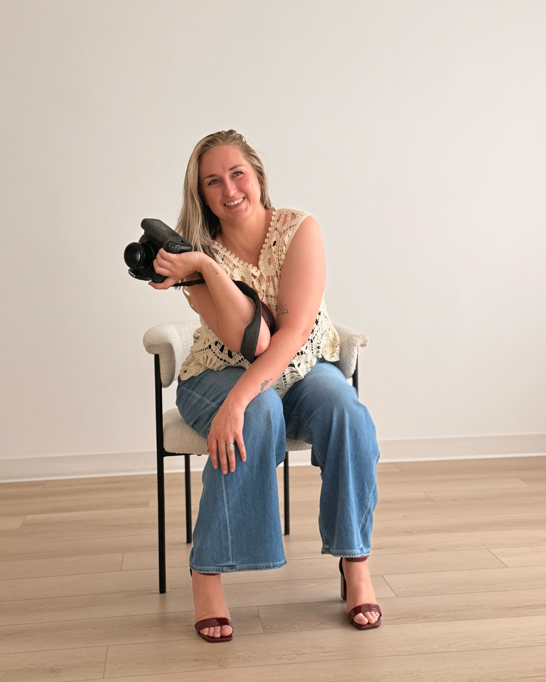 Woman with blonde hair sitting on a chair, smiling, holding a camera, wearing a beige crochet top, blue jeans, and brown heels, against a plain white wall.