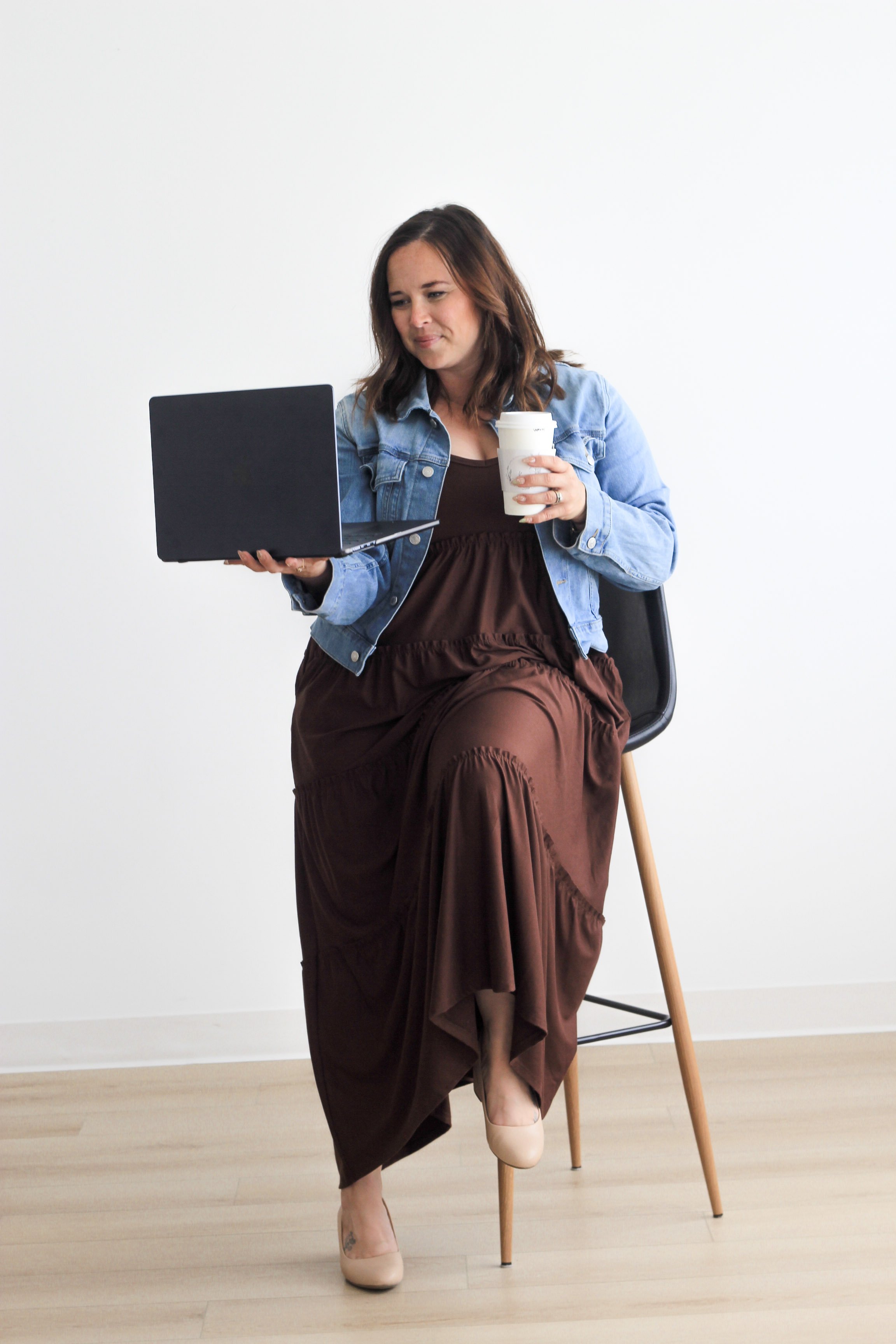 A woman sitting on a chair, dressed in a brown dress with a denim jacket, holding a small coffee cup in one hand and looking at a laptop.