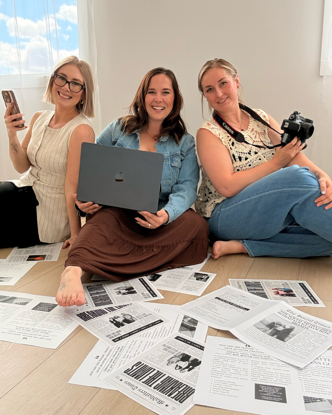 Three women sitting on the floor surrounded by scattered newspapers, holding cameras and laptops, smiling at the camera in a bright room with a window showing clouds outside.