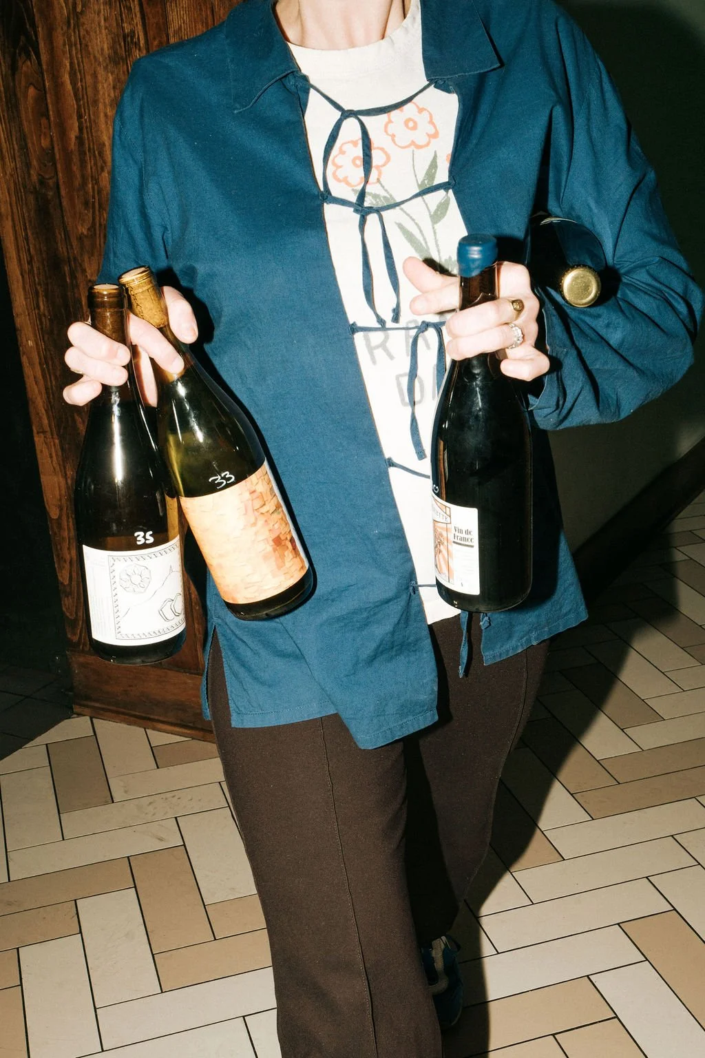 Person holding three bottles of wine, wearing a blue jacket, a floral t-shirt, and brown pants, standing indoors on a tile floor.