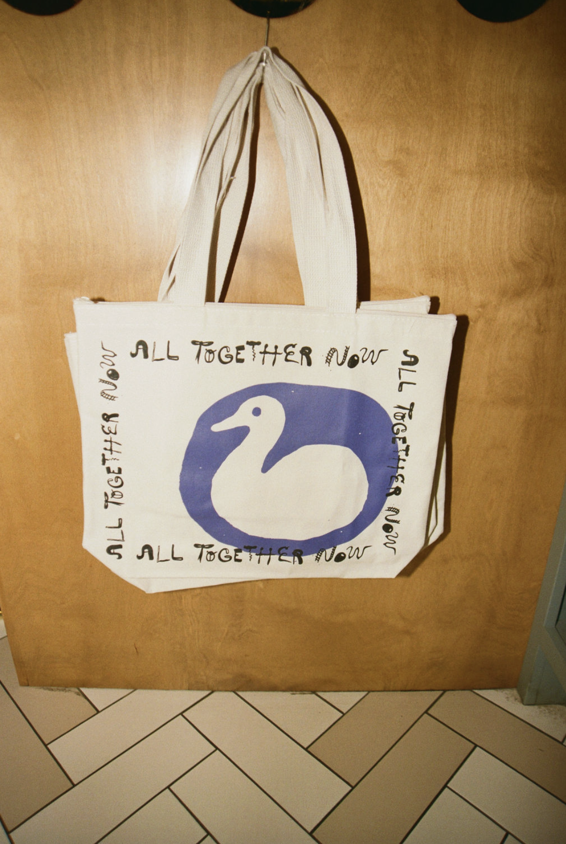 A white tote bag with a blue swan illustration and the phrase "All Together Now" written in black around the image, hanging on a wooden door with ceiling tiles above.
