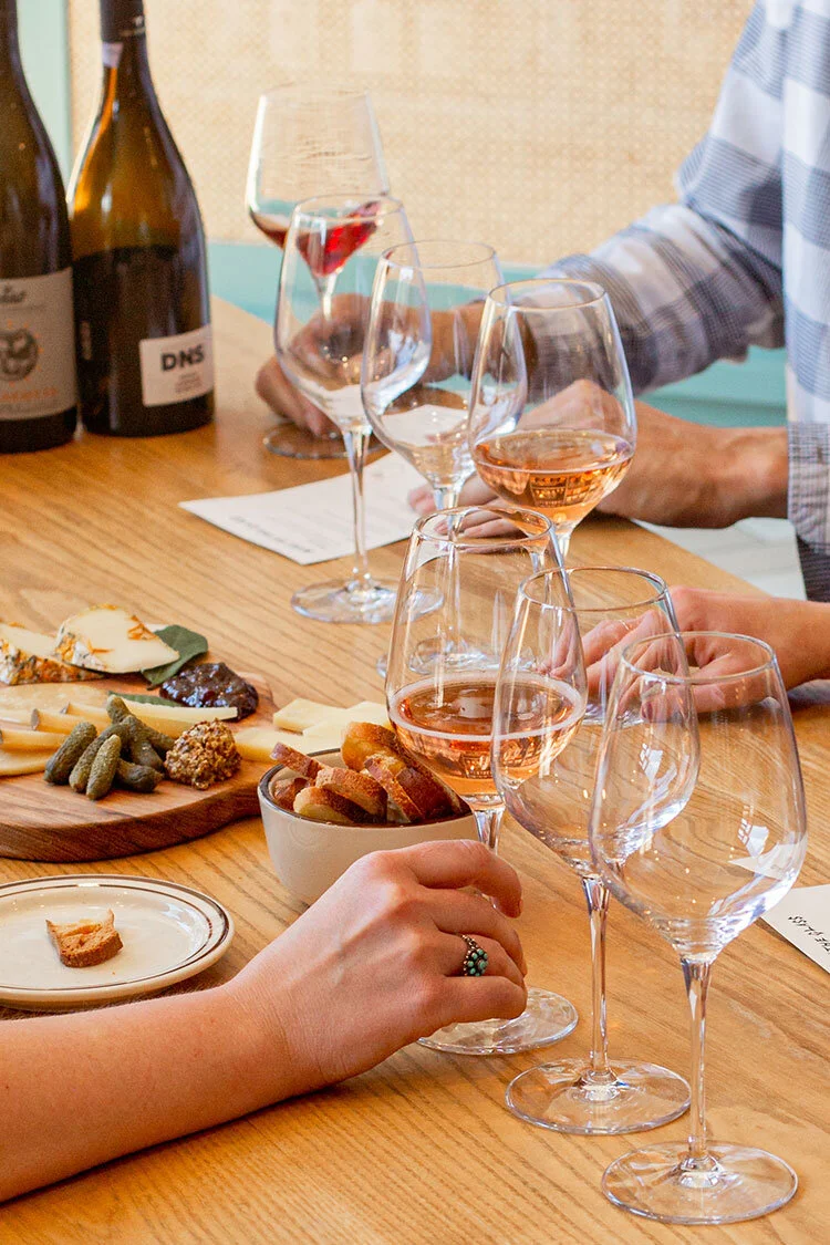 Three people sitting at a wooden table drinking glasses of wine and eating cheese at a private cheese tasting.