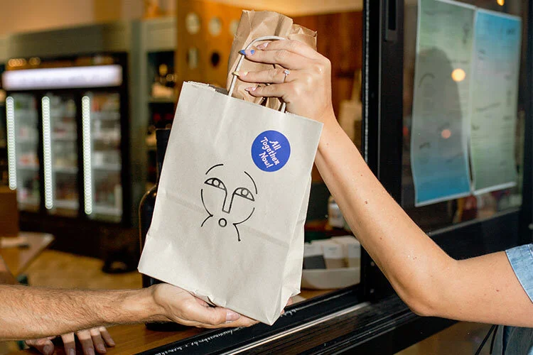 A staff member handing an All Together Now carryout bag to a guest.