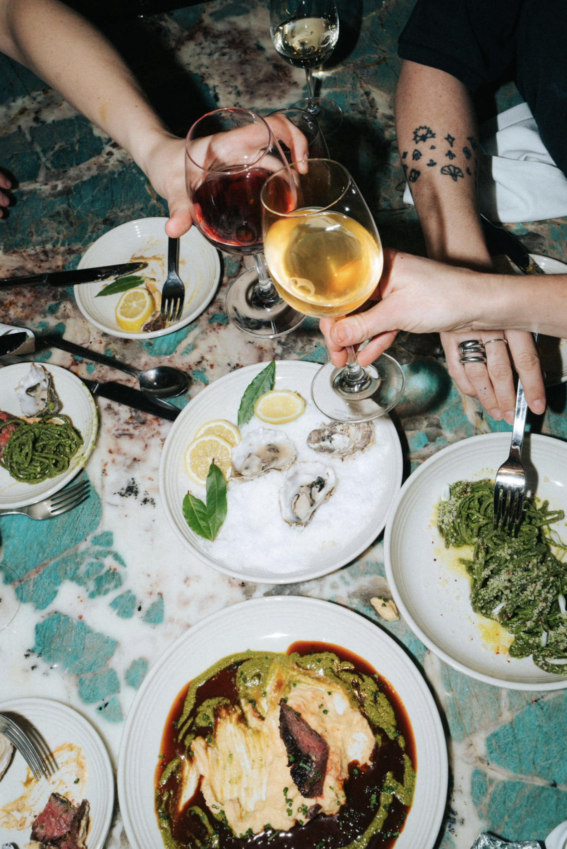 People clinking wine glasses over a table with various plates of food, including oysters, pasta, and a main dish with sauce, on a colorful marble table.