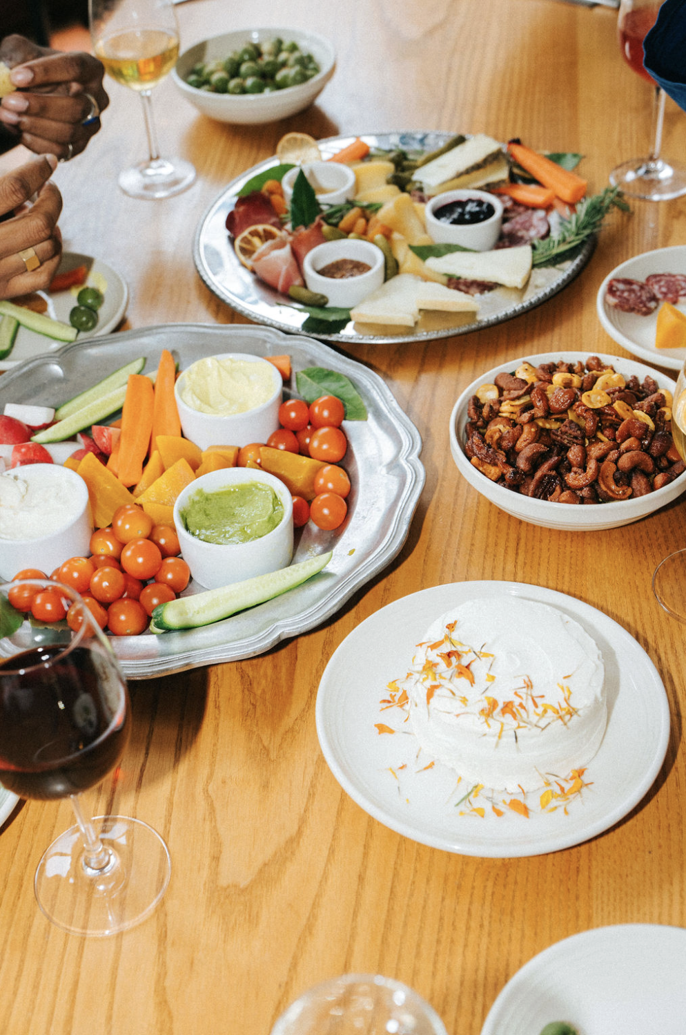 A table with various dishes including a cheese and charcuterie platter, a vegetable tray with dips, a bowl of mixed nuts, and a dessert with whipped cream and orange garnish, along with glasses of red and white wine.