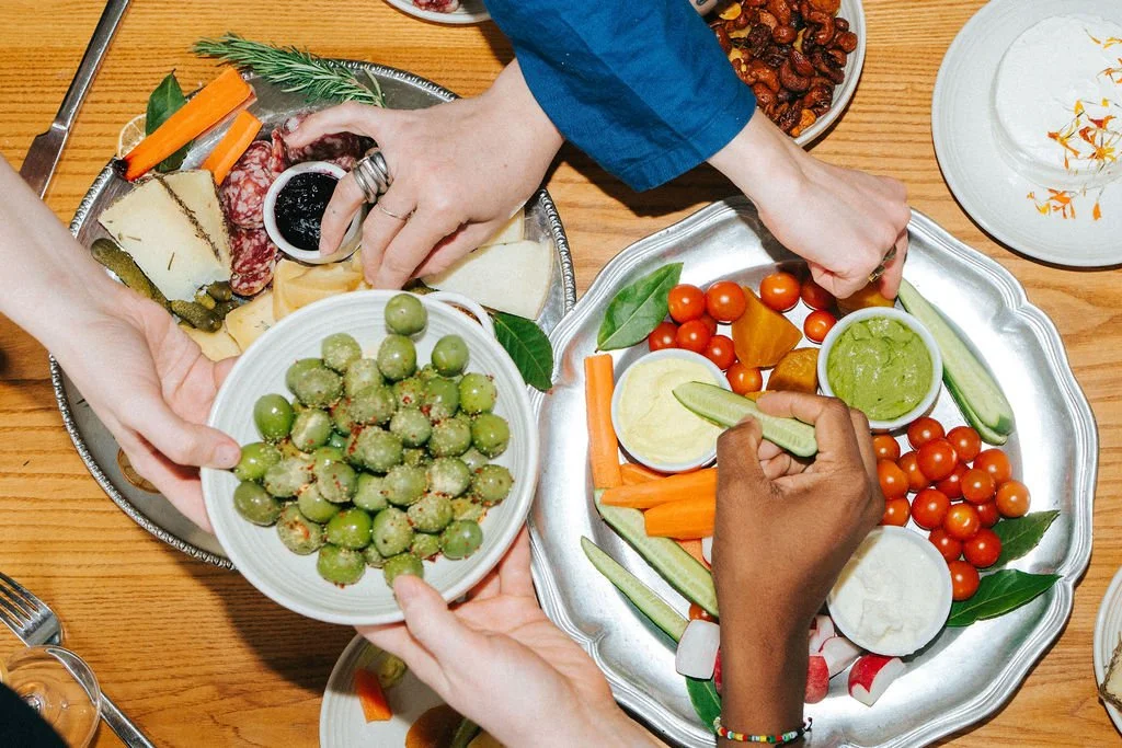 People sharing a platter of fresh vegetables, cheese, and cured meats on a wooden table.