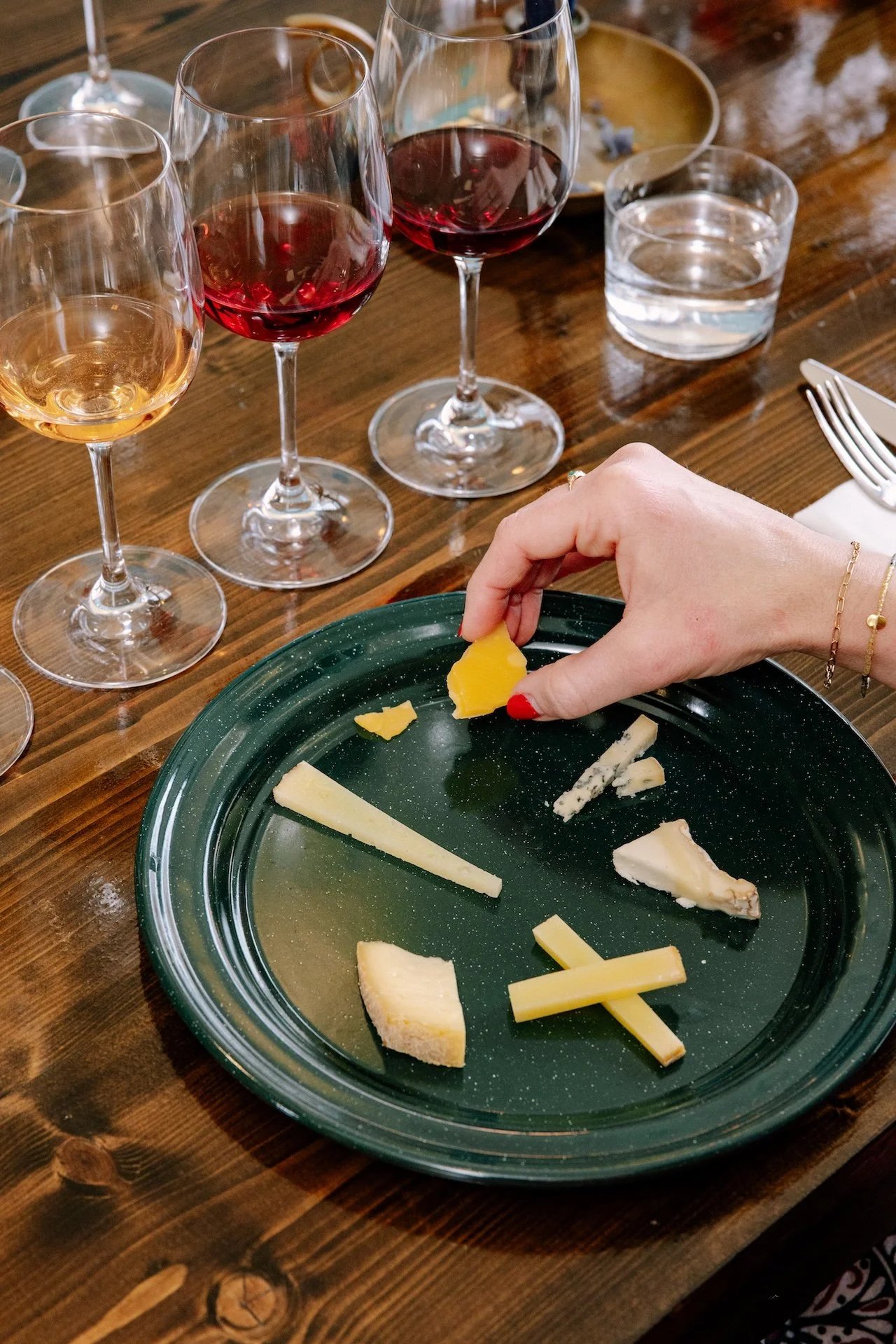 A cheese platter with various cheeses on a green round plate, a person’s hand with red painted nails picking up cheese, two glasses of red wine, one glass of white wine, a glass of water, a plate of food, a fork, and a knife on a wooden table.