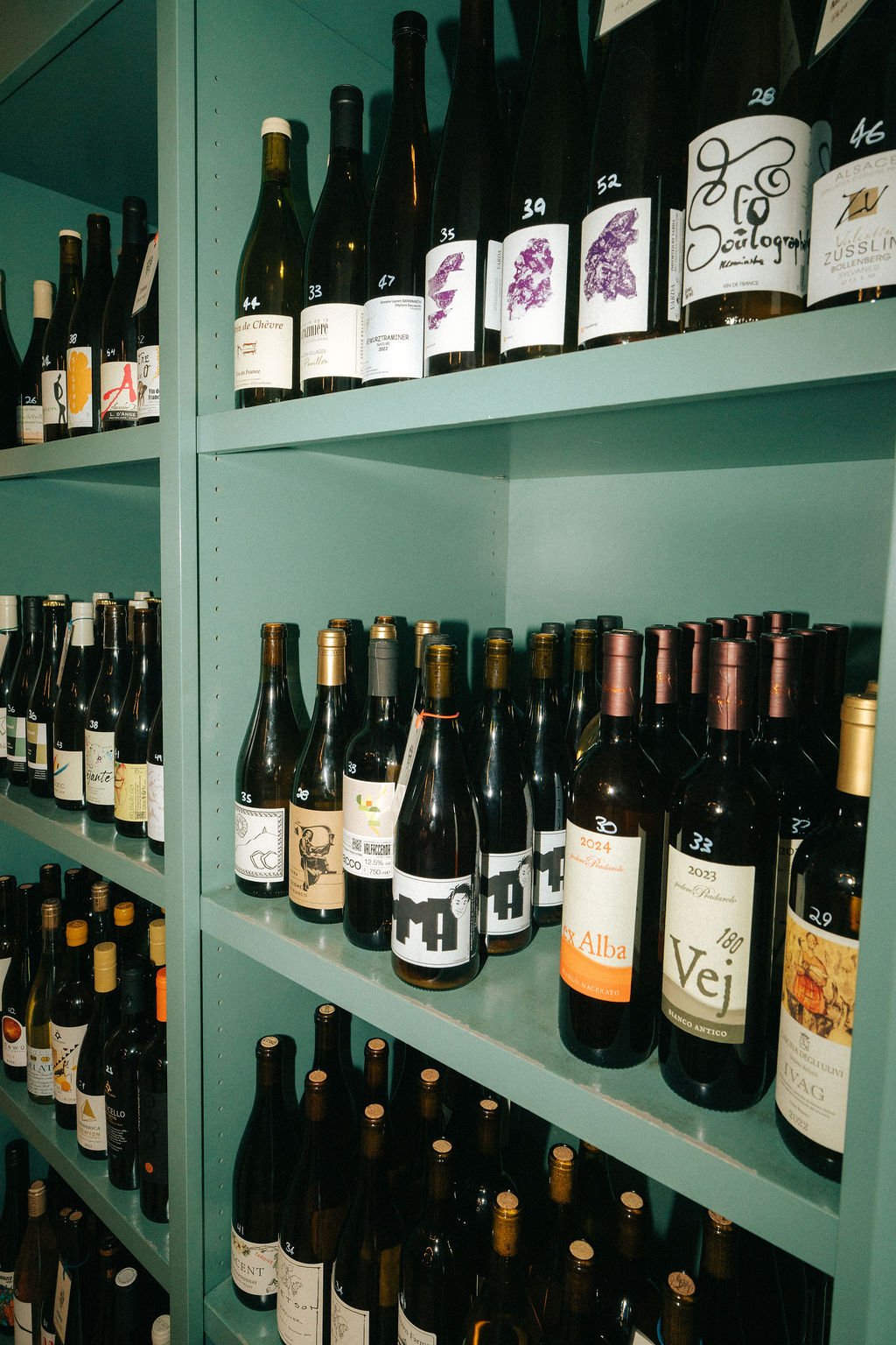 Shelf of various wine bottles with labels and caps, arranged on teal-colored shelves in a wine store.