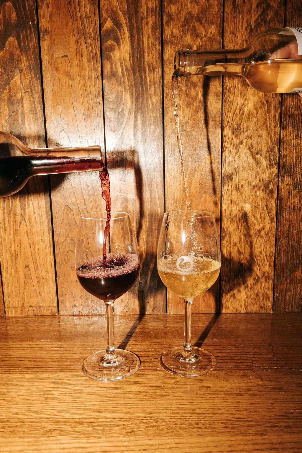 Red wine is being poured from a bottle into a glass on the left, and white wine is being poured from a bottle into a glass on the right, against a wooden background.
