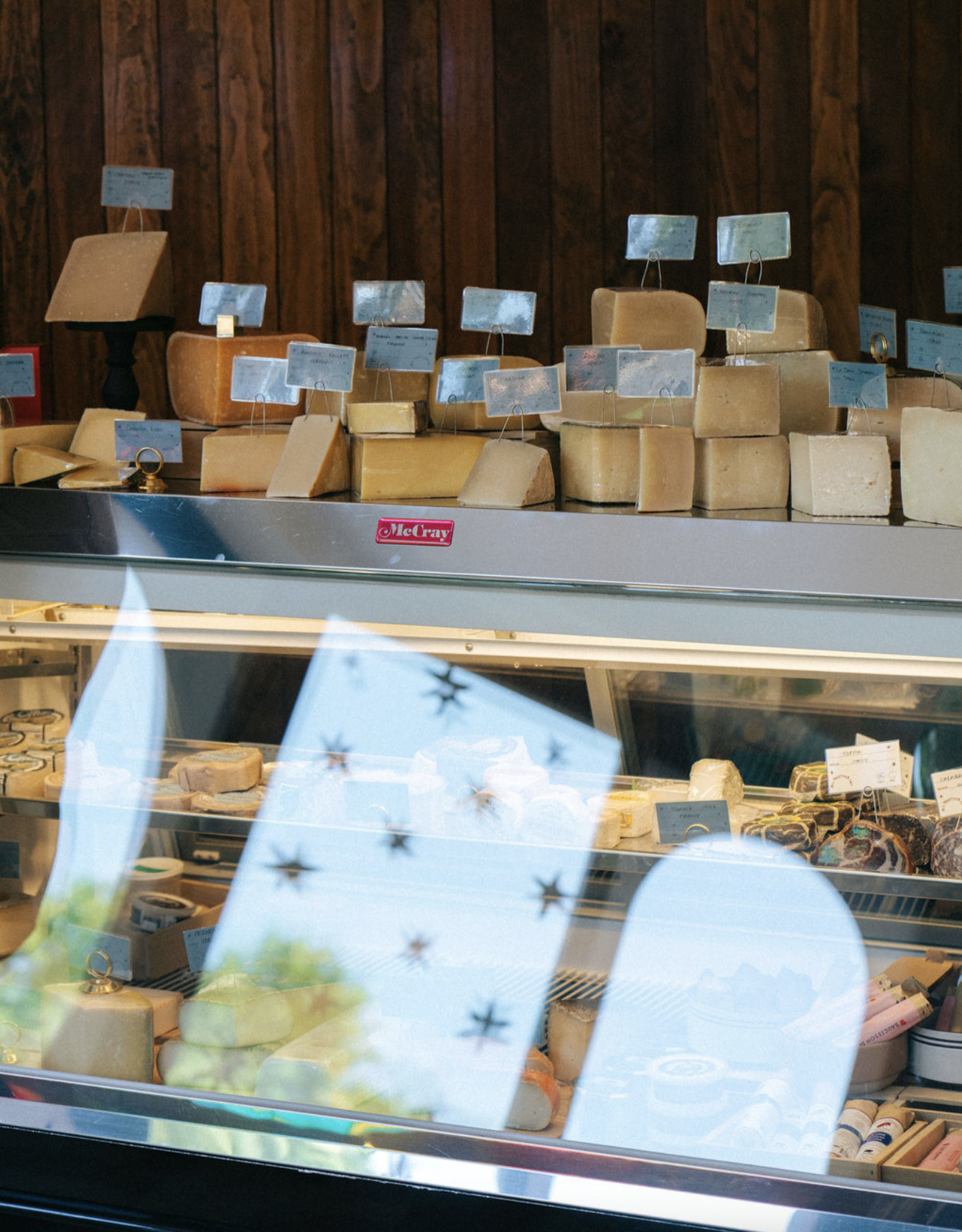 Display of various types of cheese with small signs on top, inside a shop window with a wooden wall background.