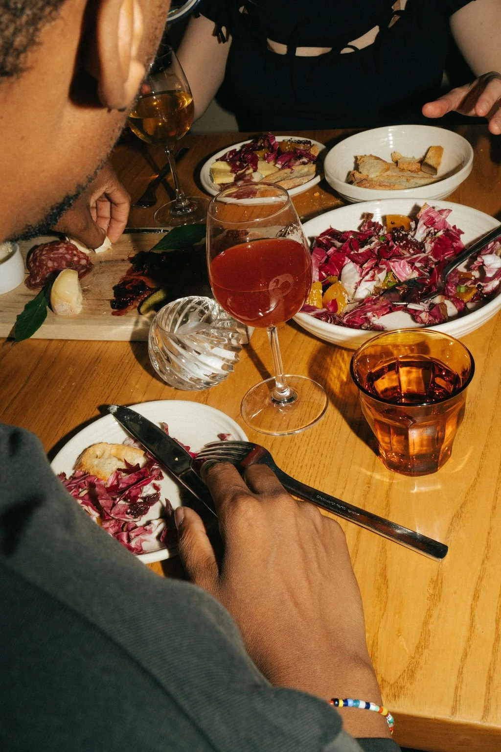 People dining at a table with various dishes, including salad, bread, and charcuterie, along with wine glasses and a decorative glass candle holder.