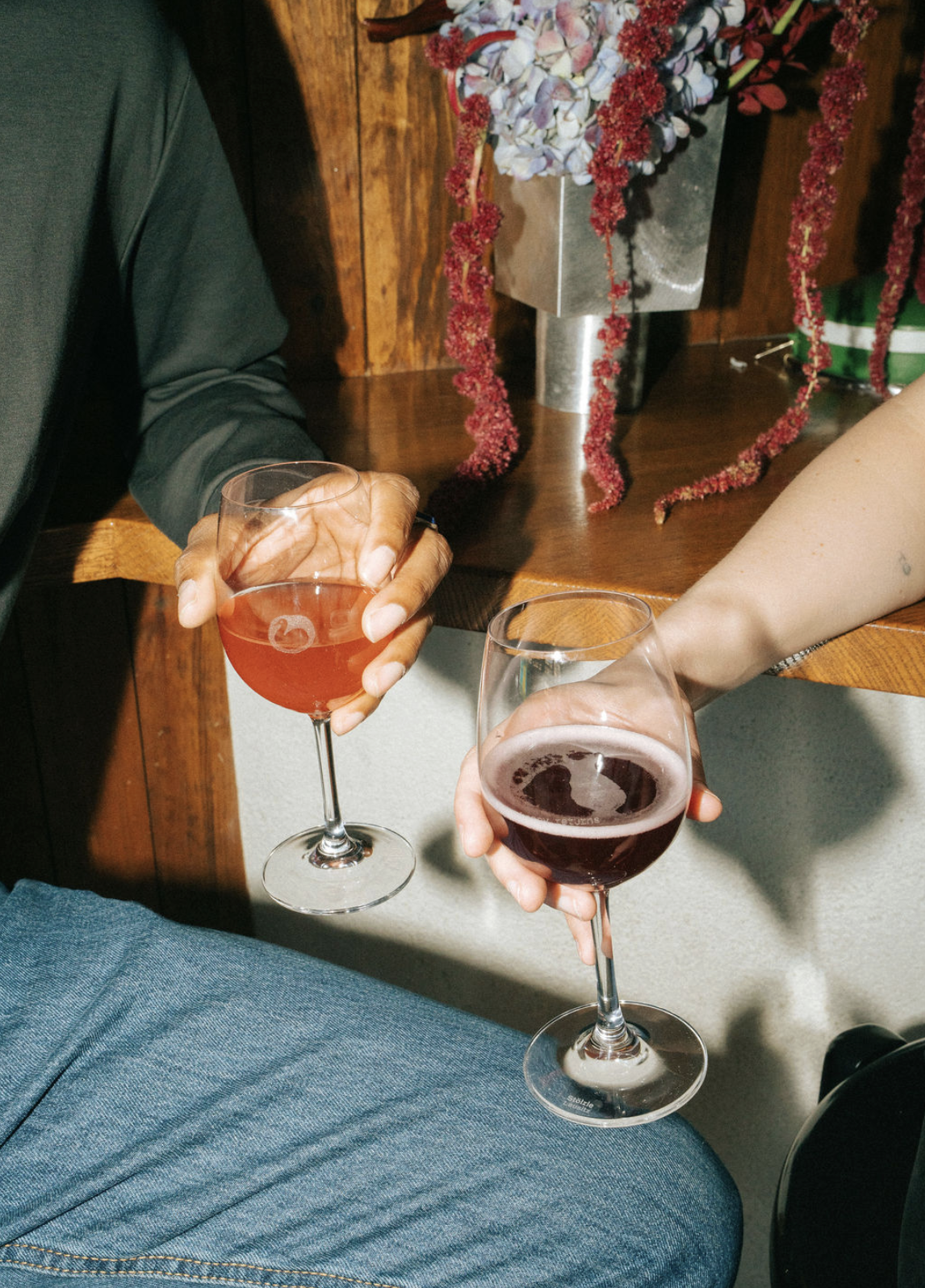 Two people holding glasses of red and pink wine during a social gathering, with a floral arrangement in the background.