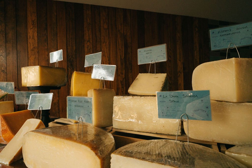 Various large blocks of cheese displayed on a wooden table with small signs indicating their types, in a cheese shop or market.