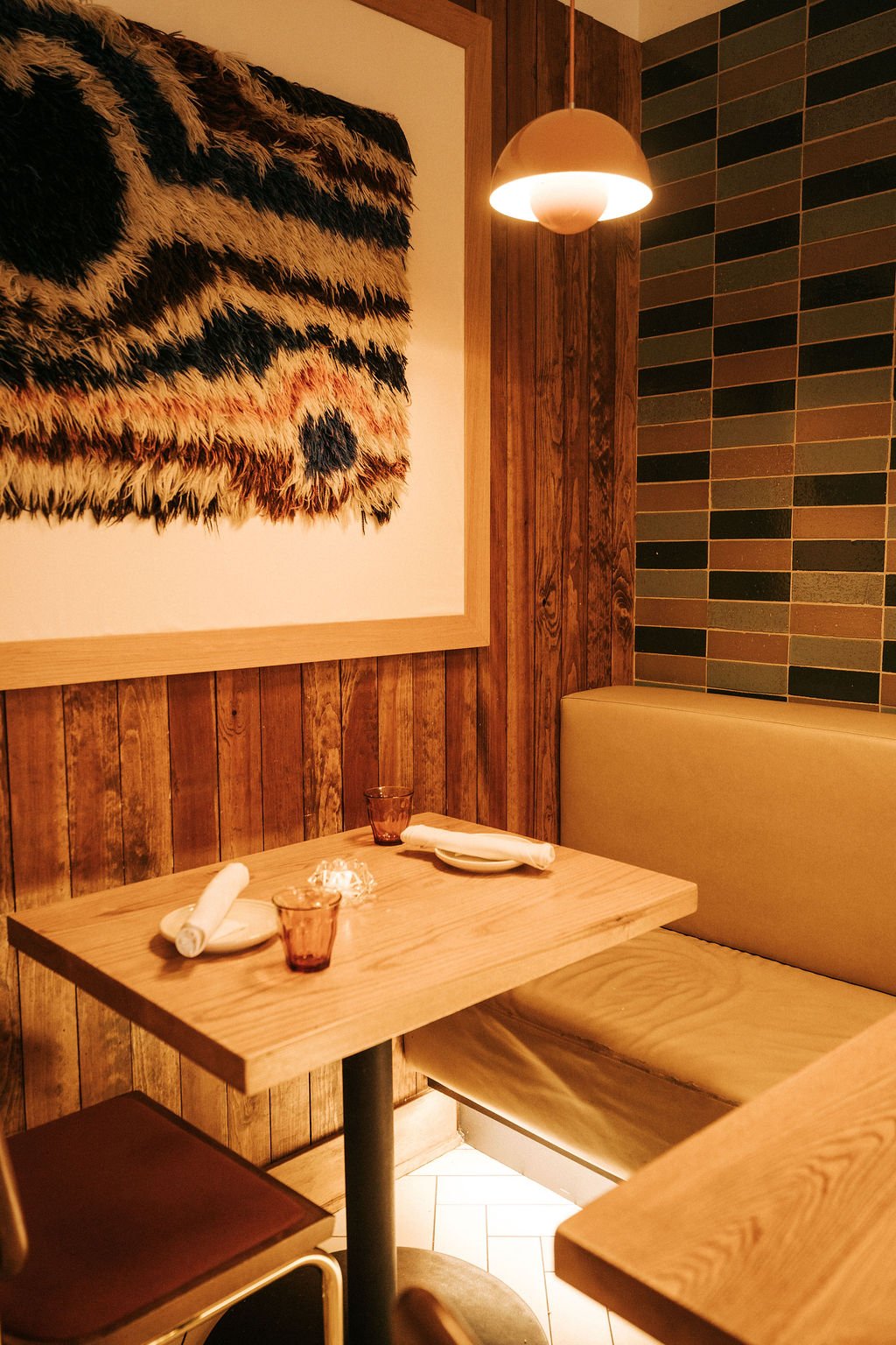 A cozy restaurant corner with a wooden table set with napkins, glasses, and plates, a bench seat, wood-paneled walls, and a large tiger print artwork on the wall illuminated by a hanging pendant light.