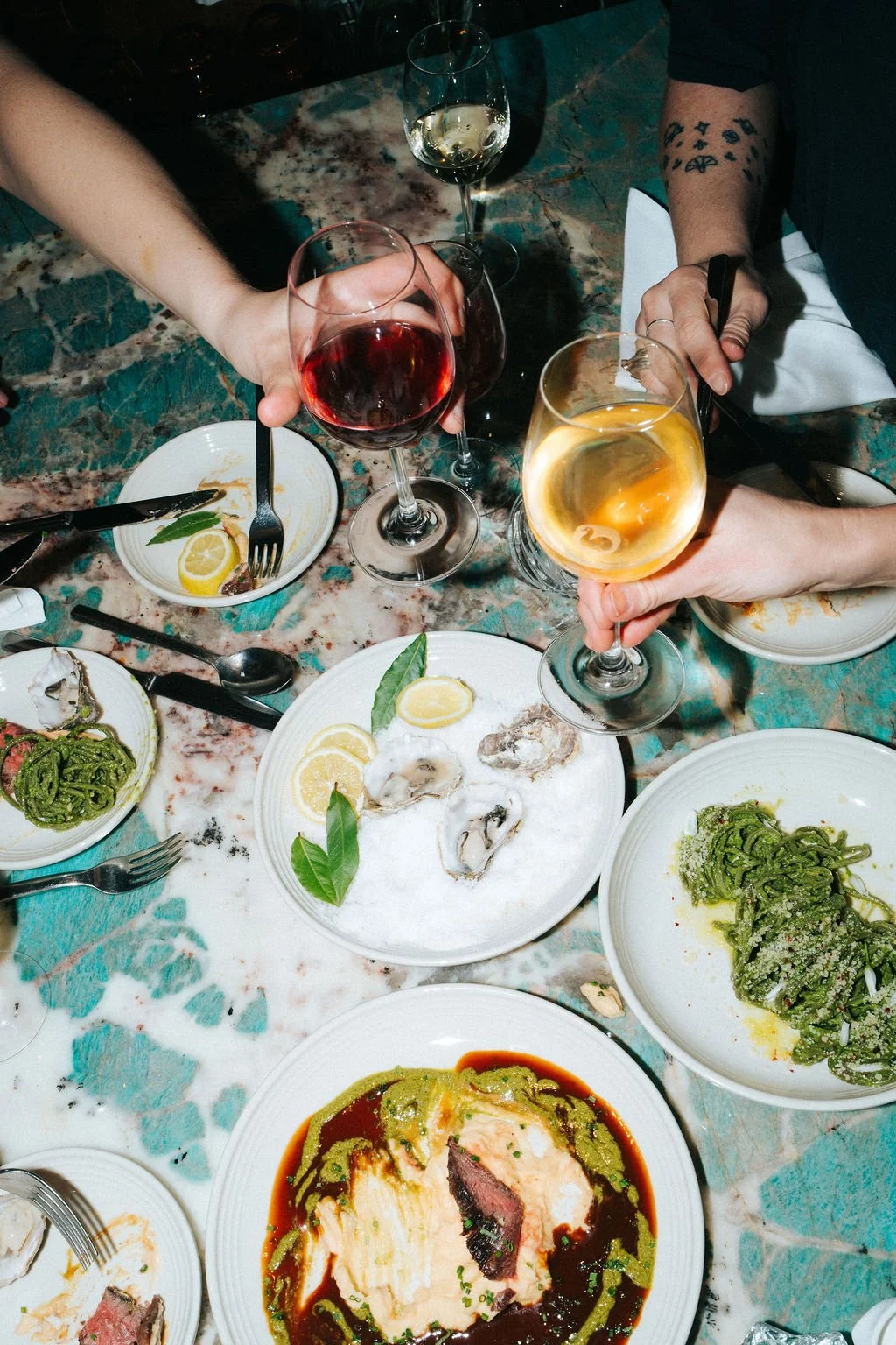 People clinking glasses of white wine and red wine at a dinner table with plates of oysters, pasta, and steak, on a marble bar.