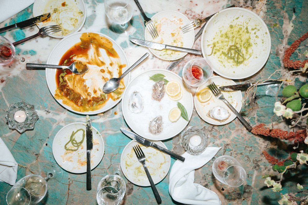 Empty and partially filled plates, glasses, and utensils on a table with leftover food and drinks, indicating a recently finished meal at a social gathering.