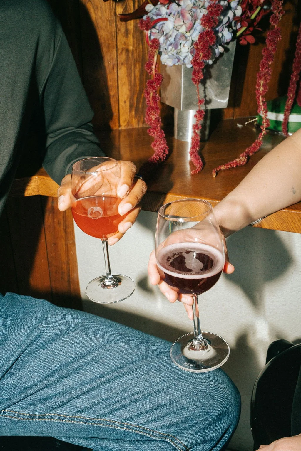 Two people clinking glasses of red and pink wine in a cozy indoor setting with wooden furniture and floral decorations.