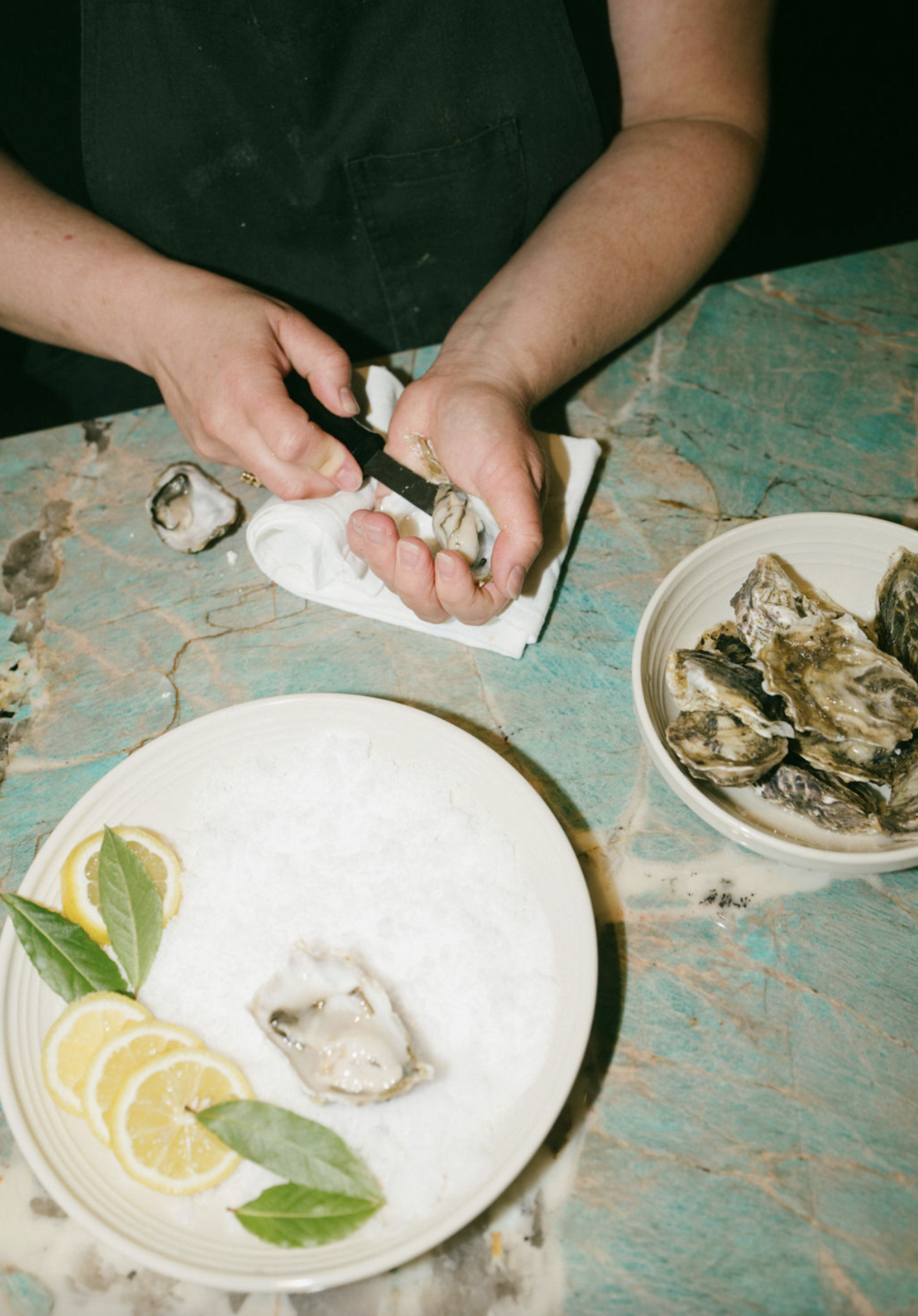 Someone shucking oysters on a blue-green marble countertop, with a white plate with lemon slices and green leaves, and a bowl of oysters nearby.