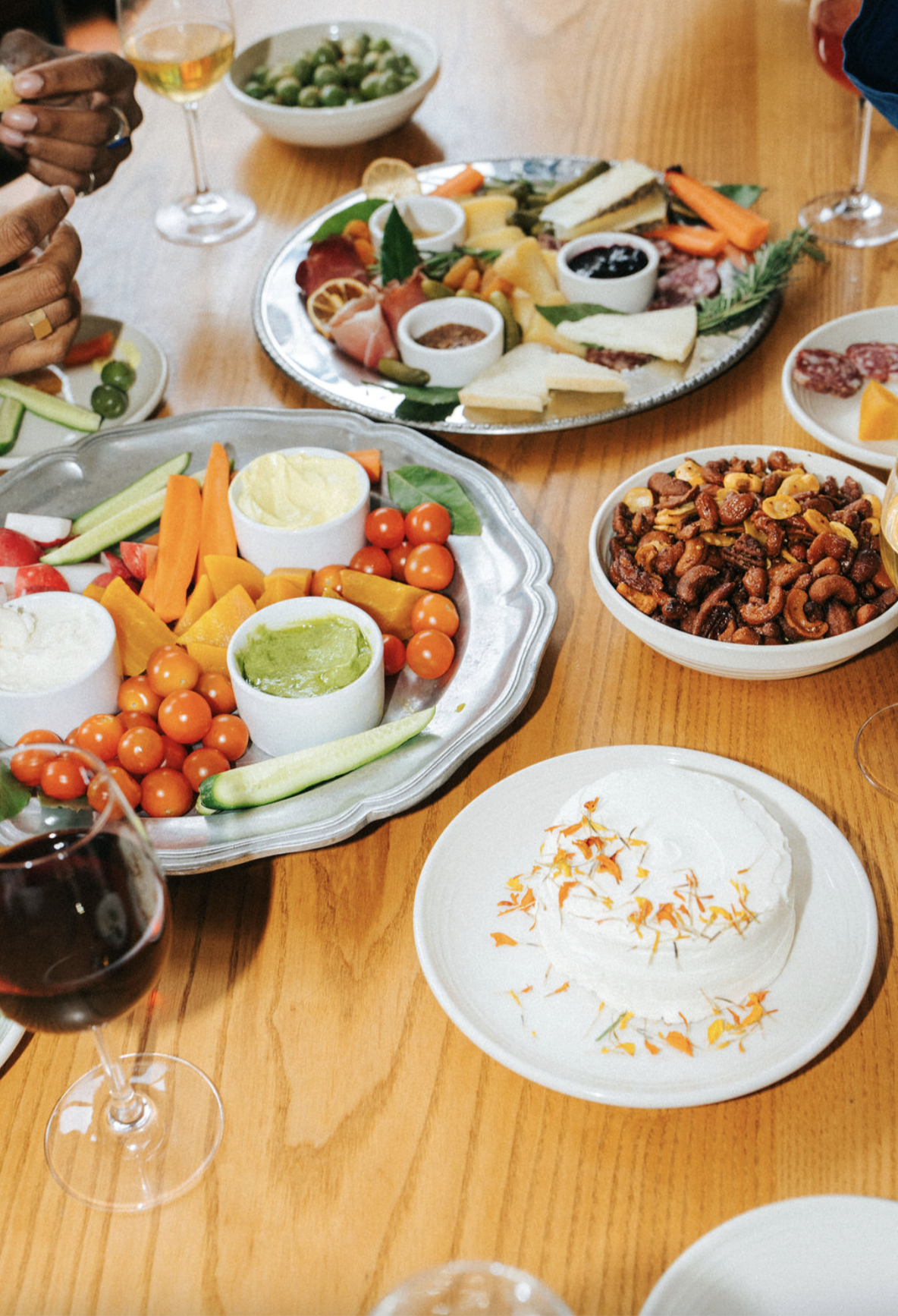 A varied charcuterie and vegetable platter, bowls of dip, a bowl of mixed nuts, and glasses of red and white wine on a wooden table.