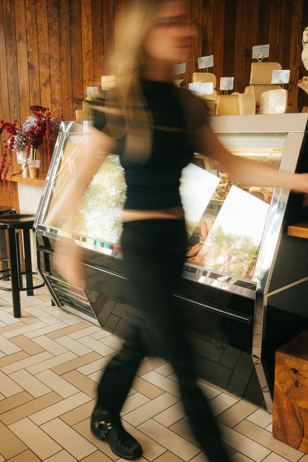 A woman wearing black clothing is walking past a cheese case in a restaurant with wood-paneled walls and stools in the background.