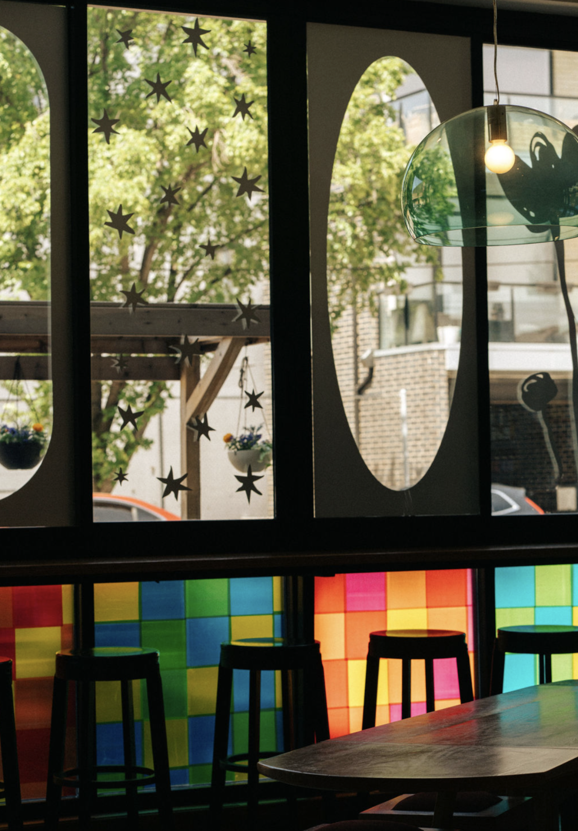Inside a colorful cafe or restaurant with a window view of trees outside. The window has star-shaped decorations. There is a hanging light fixture with a bulb. In the foreground, there's a wooden table and four colorful stools with illuminated multicolored blocks on the wall behind them.