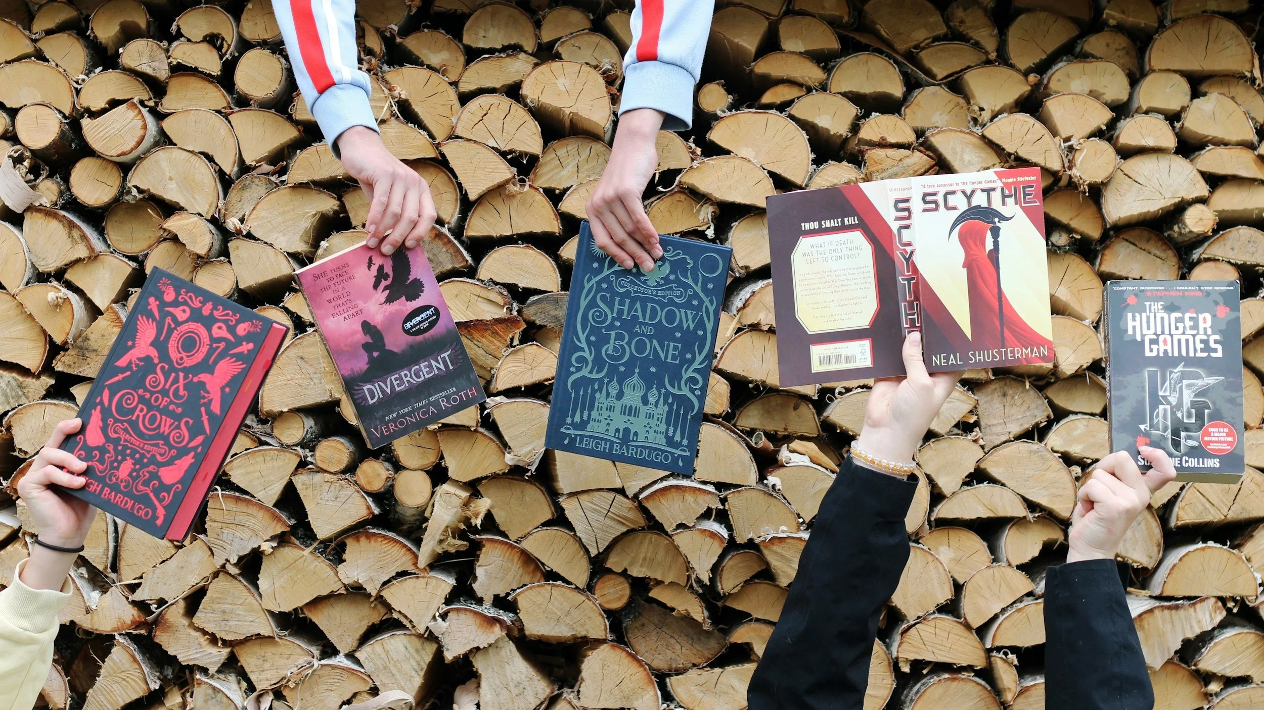 People holding various books in front of a large stack of cut firewood. The books include 'A Song of Crows,' 'Divergent,' 'Shadow and Bone,' 'Thou Shalt Kill,' 'The Hunger Games,' and others. The hands are visible, but the faces are not shown.